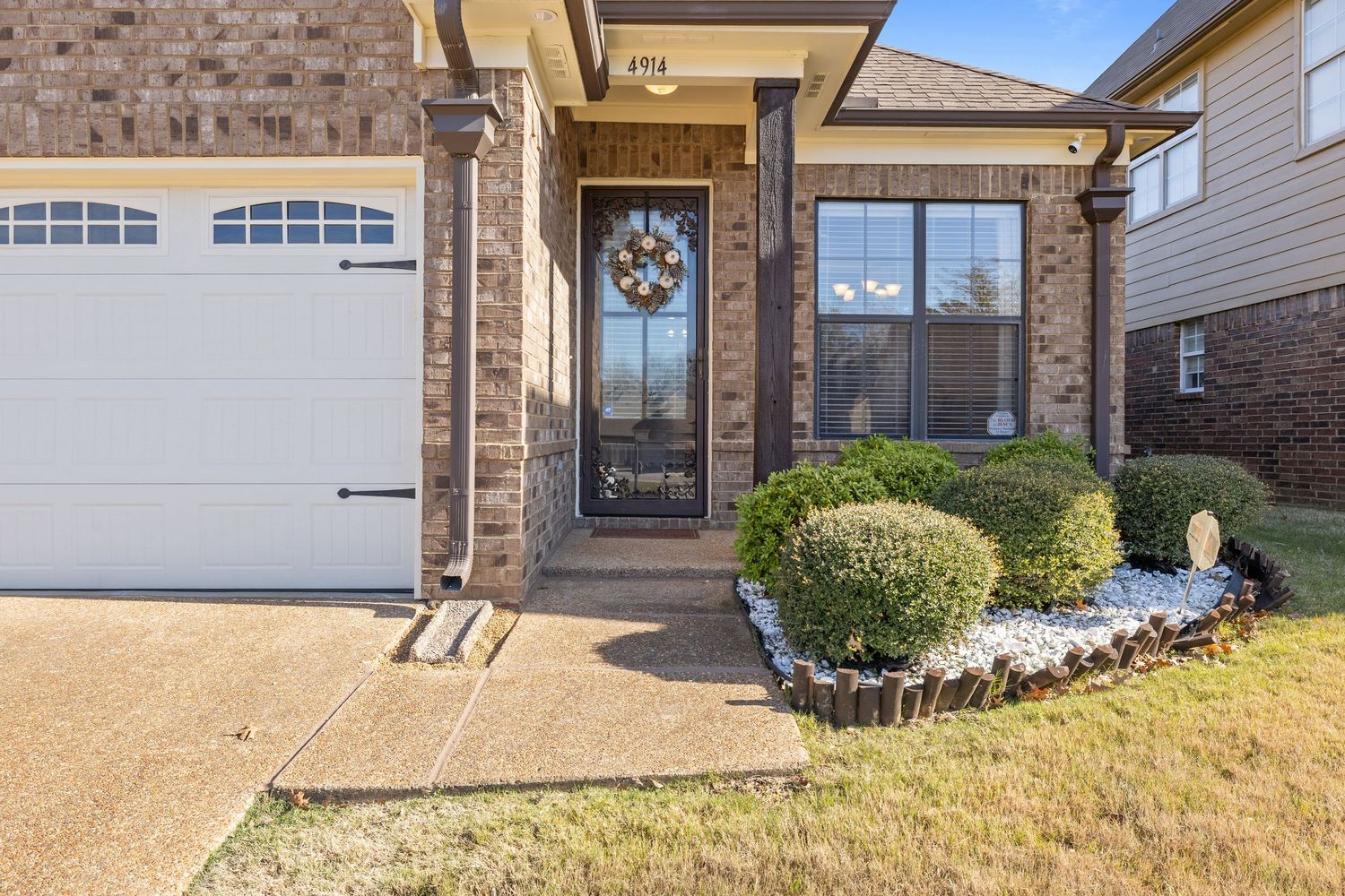 Front entrance of a brick house with white garage door, decorative wreath on front door, and manicured shrubs along walkway.