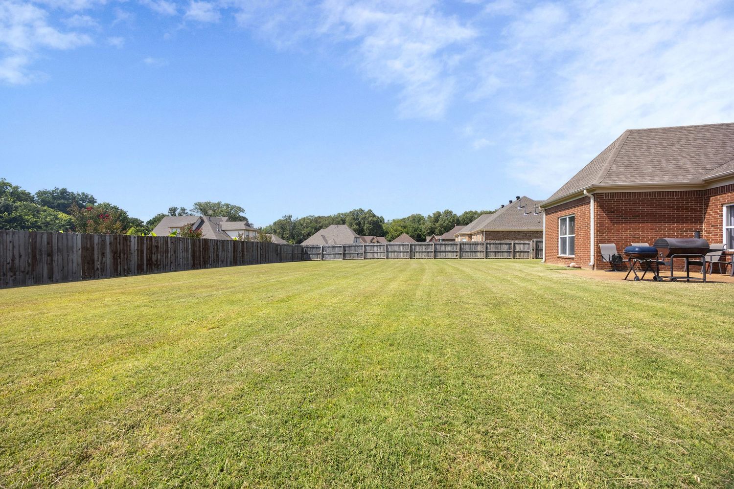 Large well-maintained backyard with green lawn, wooden fence, and brick house with patio visible in background.