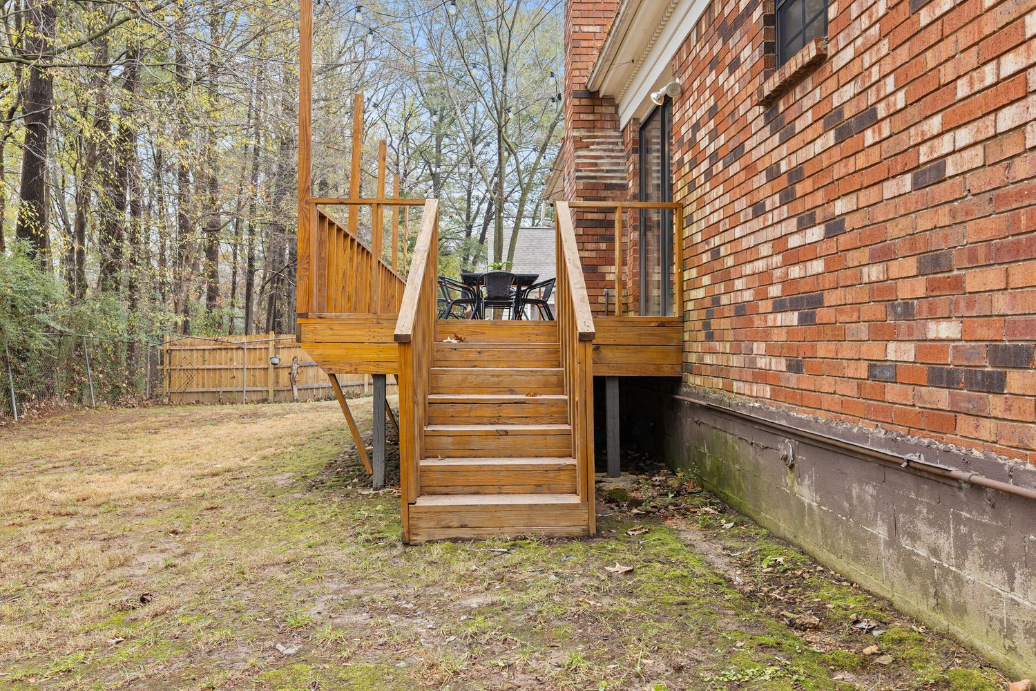 Exterior view of brick house with wooden deck and stairs surrounded by trees.