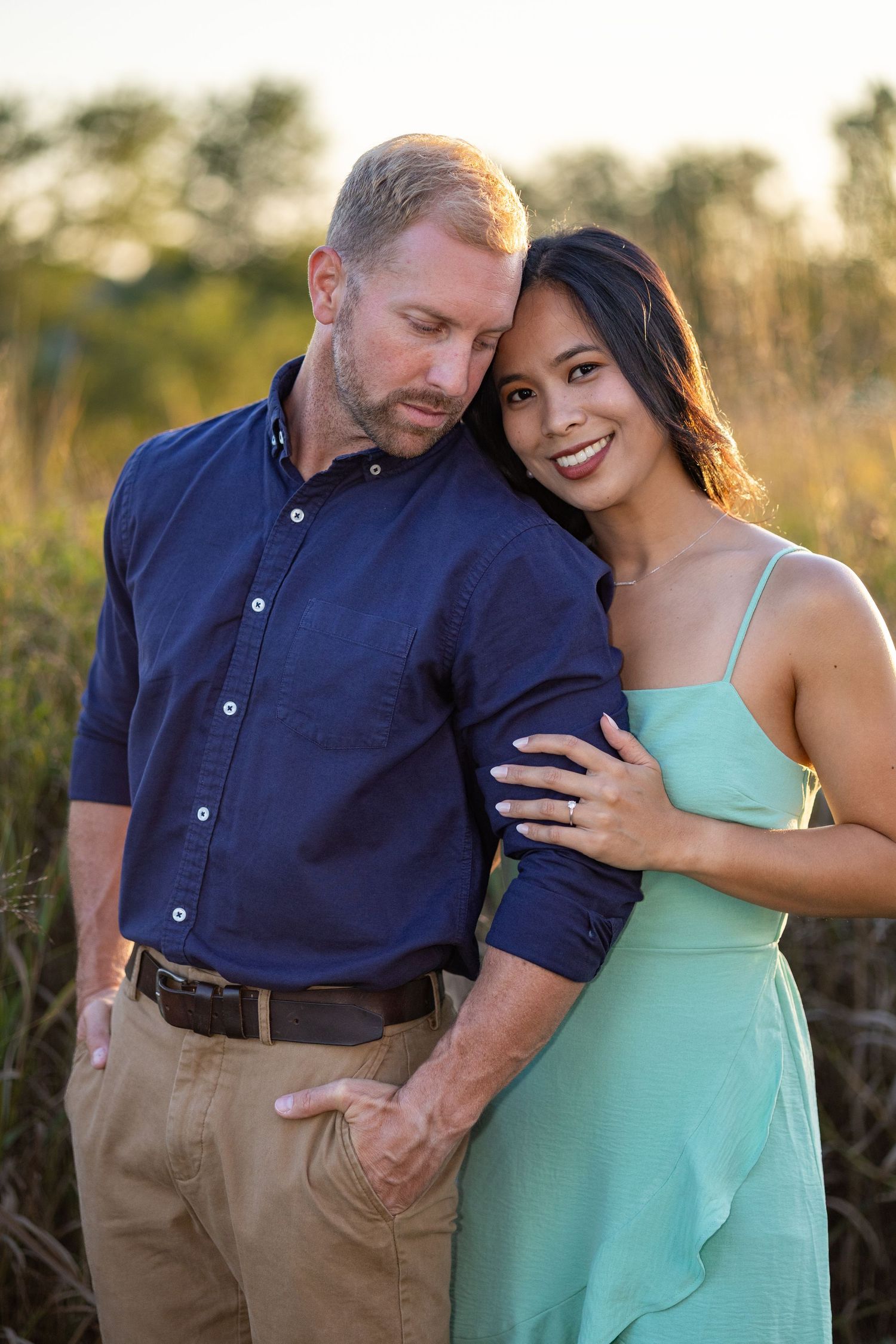 A romantic couple poses together in tall grass at sunset, she in turquoise dress and he in navy button-down.