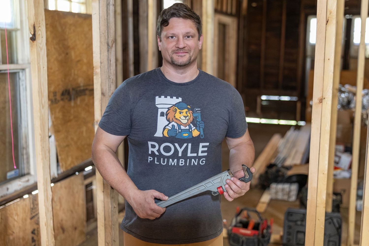 Plumber in Royle Plumbing t-shirt holding a handsaw inside a wood-framed construction site.
