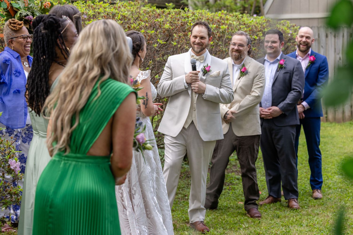 Two grooms in suits laugh with wedding guests outdoors during a joyful garden wedding ceremony.