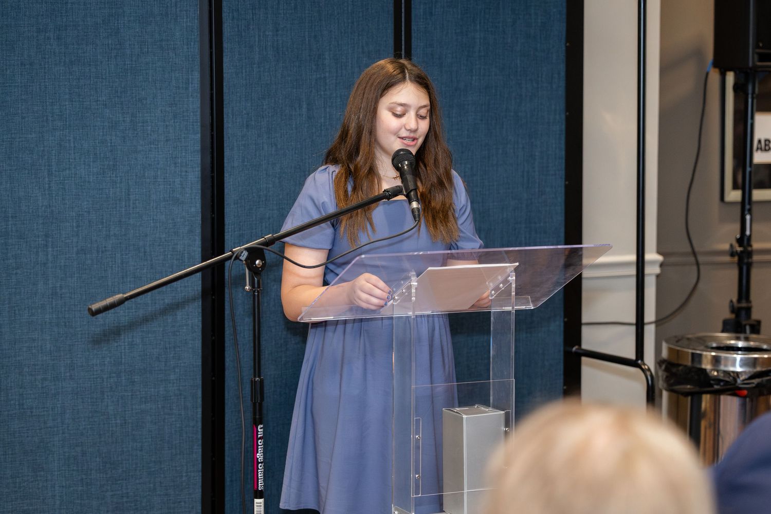 Young girl in blue dress speaking at a clear podium with a microphone at a formal event.