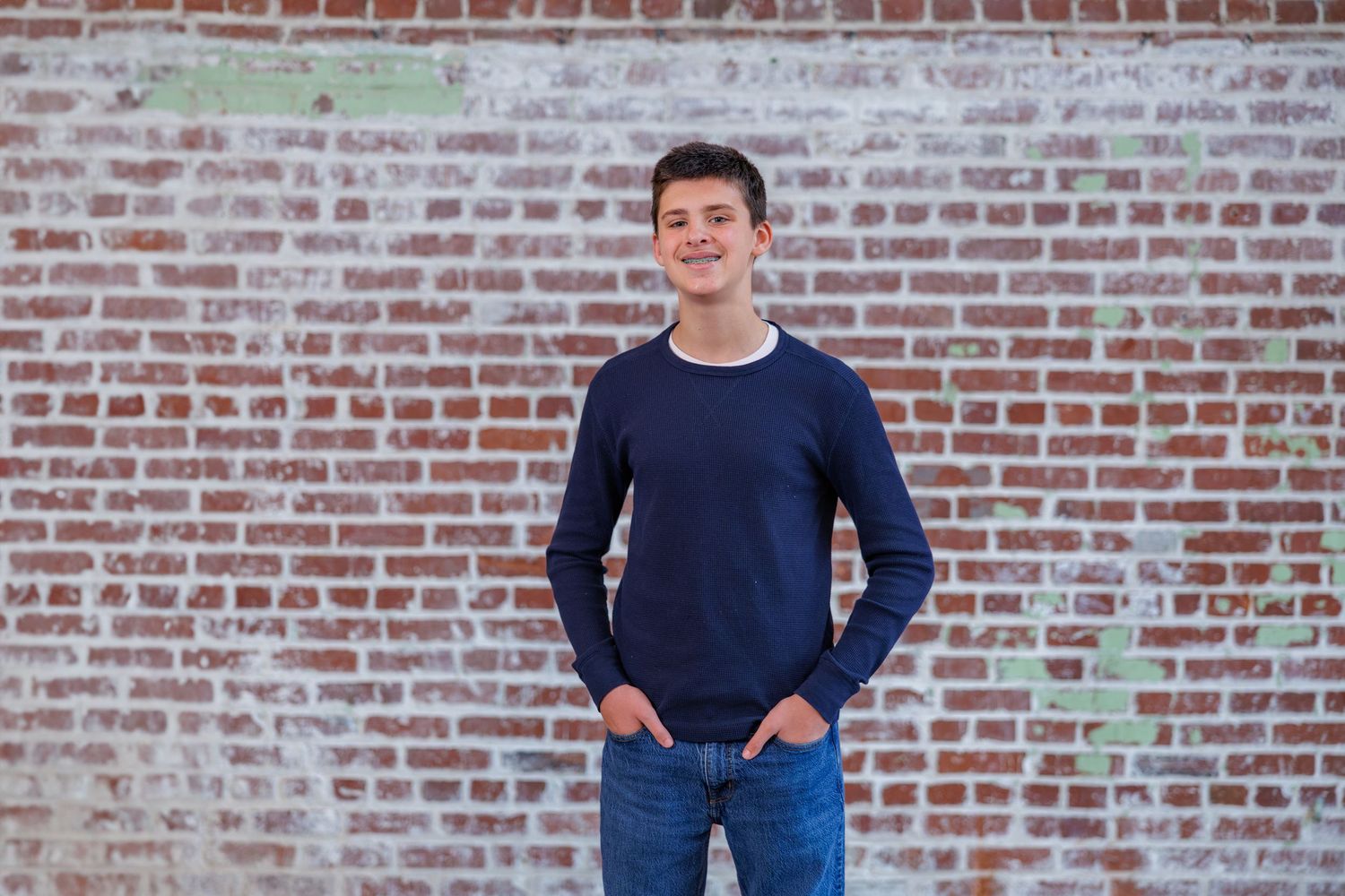 A person in a navy sweater and jeans stands casually against a rustic brick wall background.