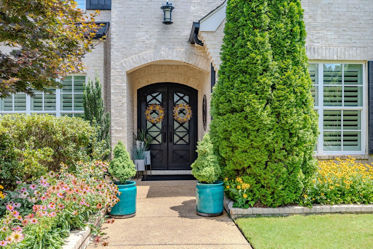 A luxurious residential entrance with white brick, green shutters, and potted plants flanking the black front door.