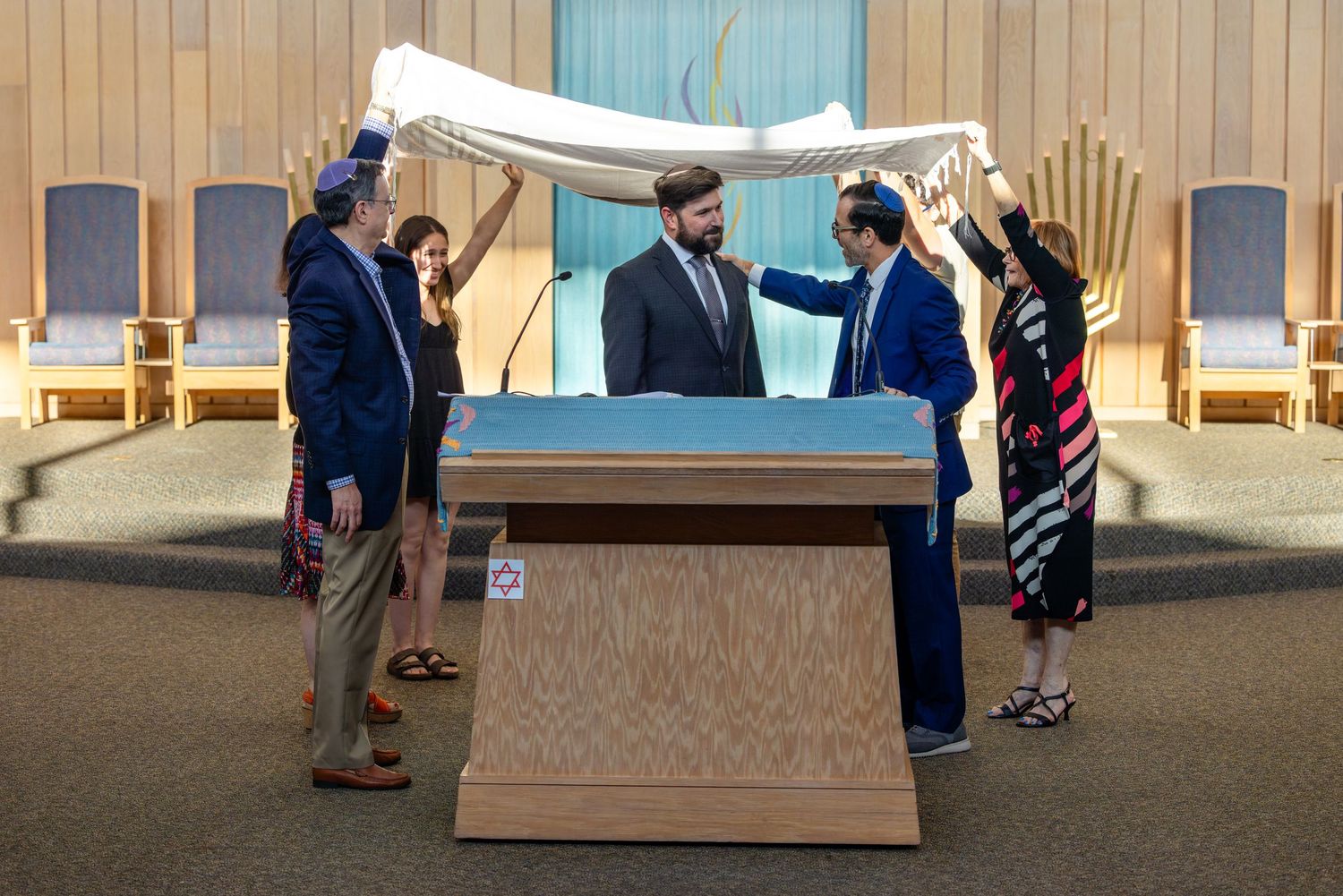 Jewish wedding ceremony under a chuppah in a synagogue with four people holding the canopy over the couple.