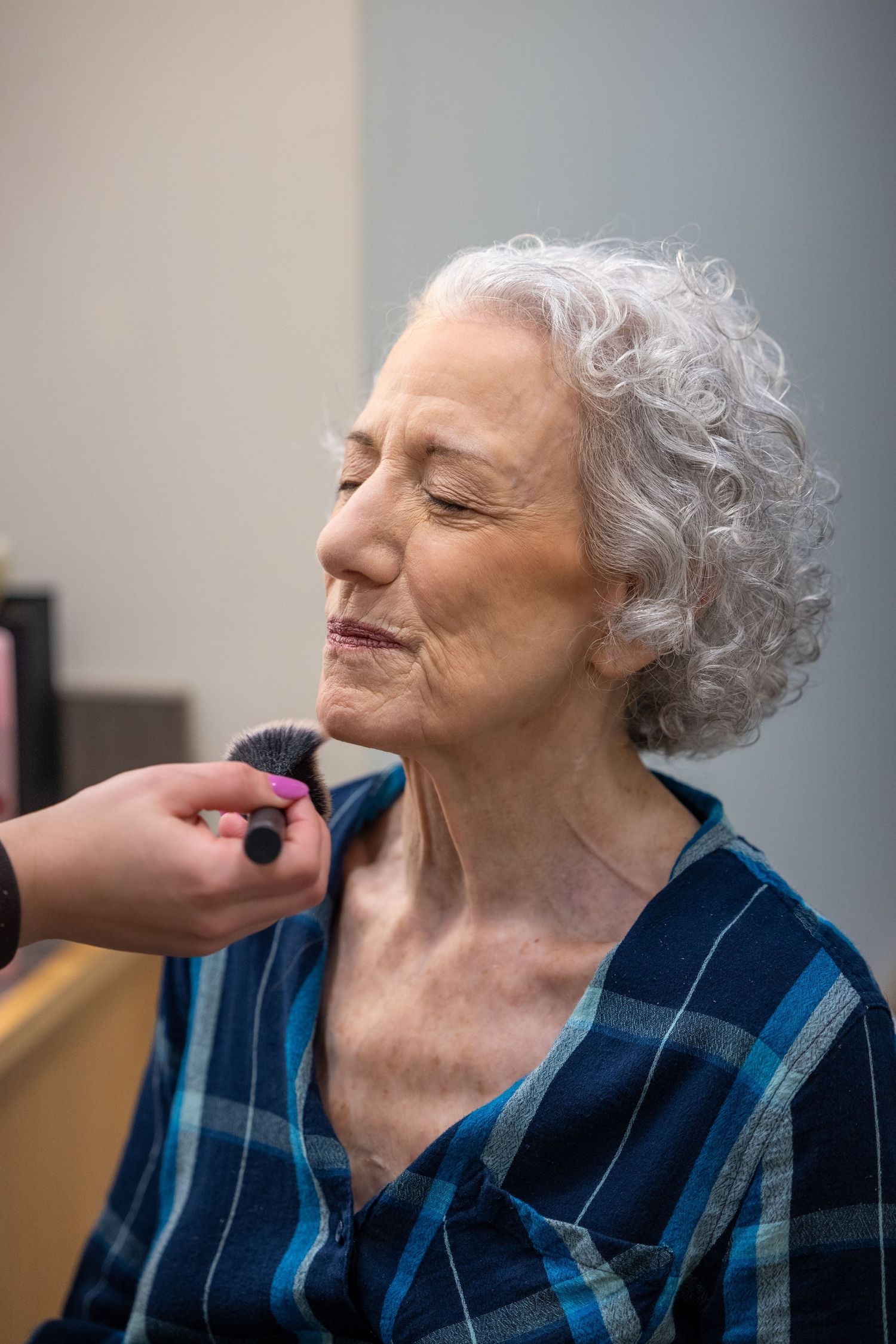 An elderly person with silver curly hair and closed eyes wearing a blue plaid shirt receives care from someone's hand.