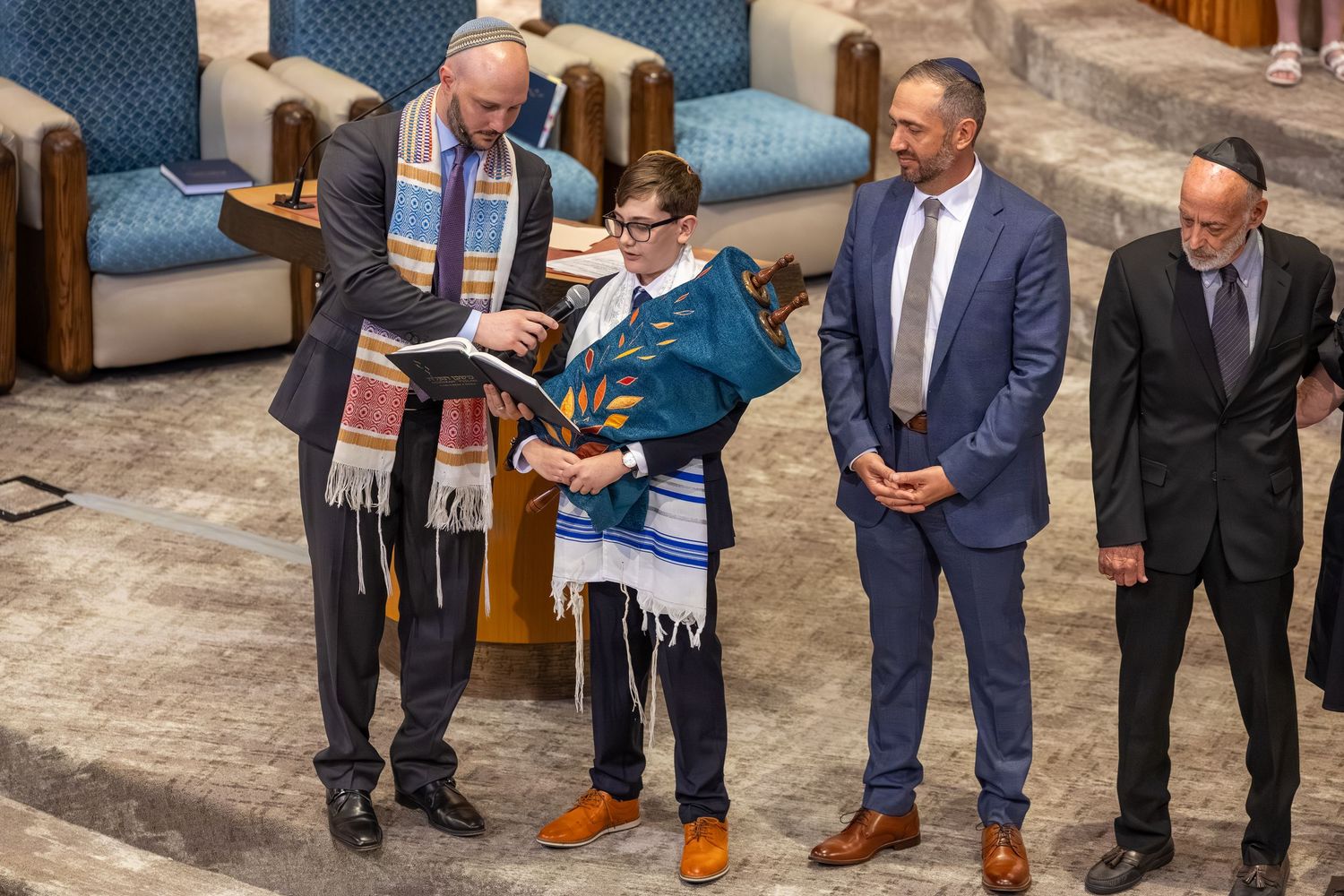 Young boy in tallit reads from Torah during Bar Mitzvah ceremony with rabbi and family members standing nearby.