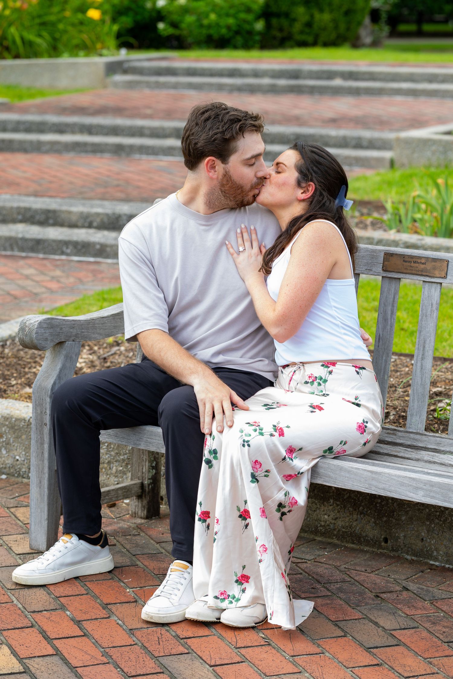 Couple sharing a romantic kiss on a wooden park bench, surrounded by lush greenery and brick pavement.
