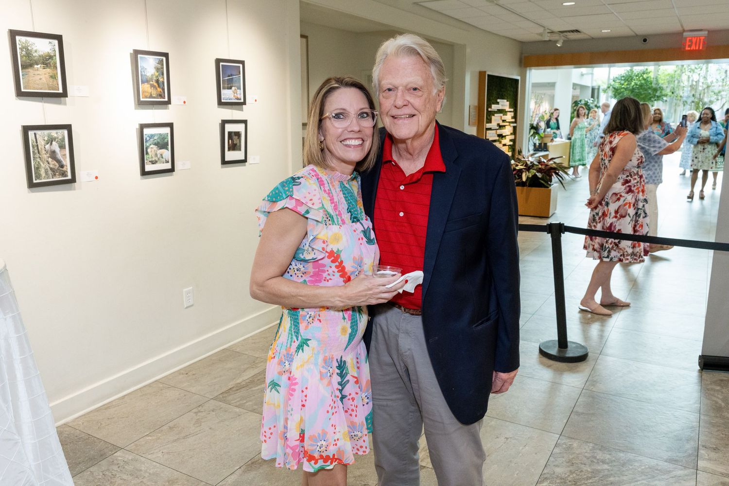 A smiling couple poses together at an art gallery event, with framed artwork displayed on white walls behind them.