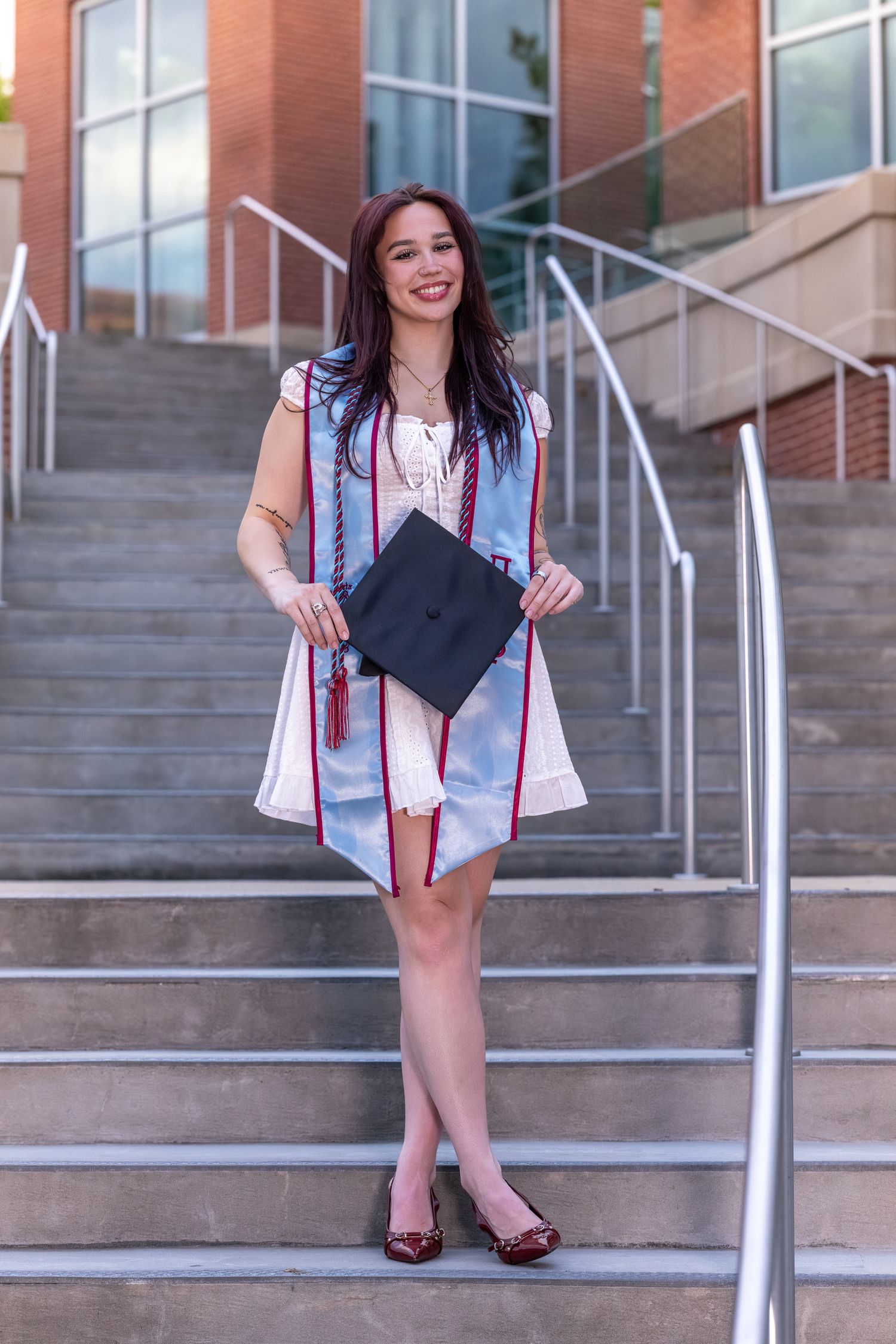Smiling young woman in graduation gown and cap posing on university steps, celebrating her academic achievement.