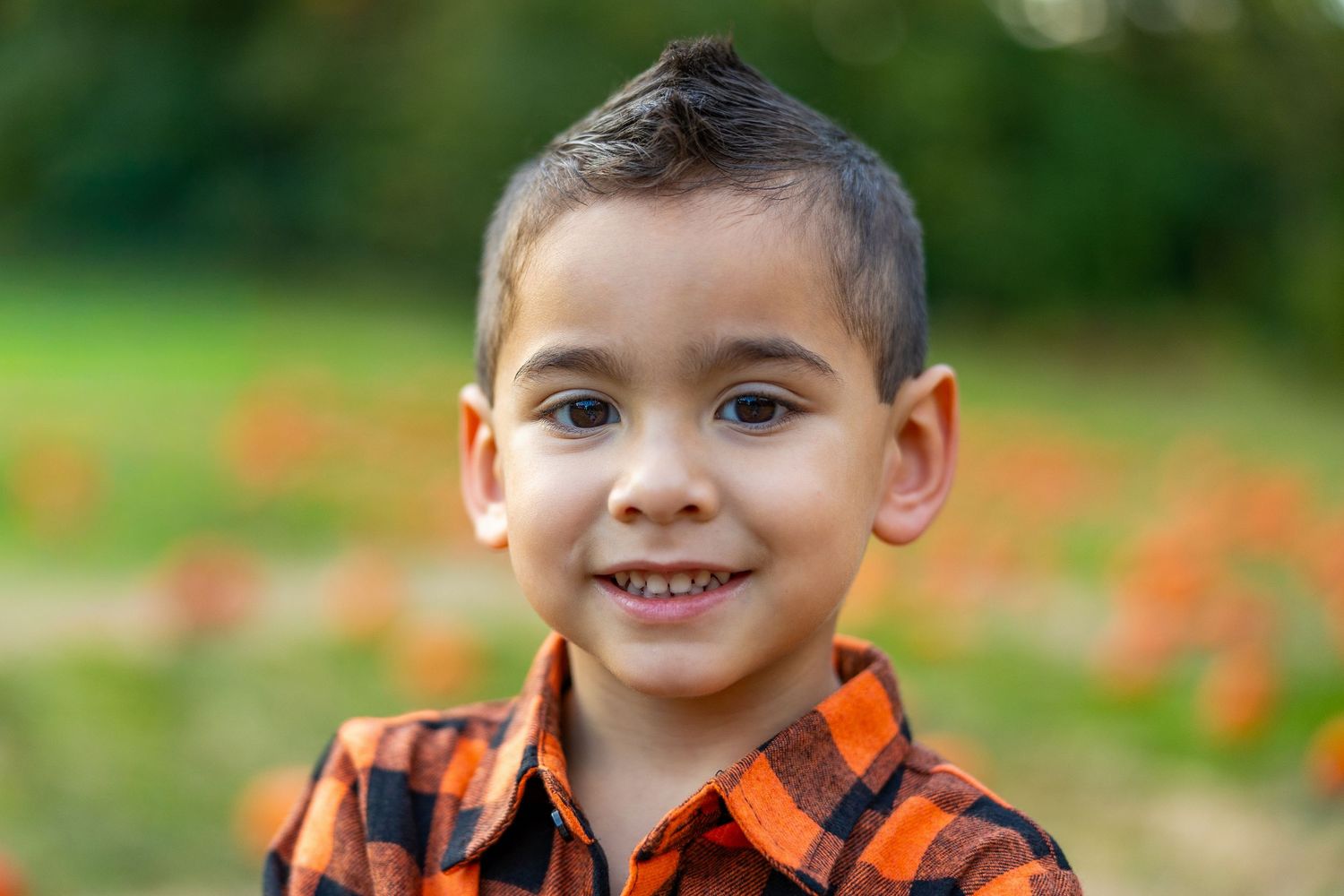Child wearing orange plaid shirt smiles brightly in pumpkin patch setting.