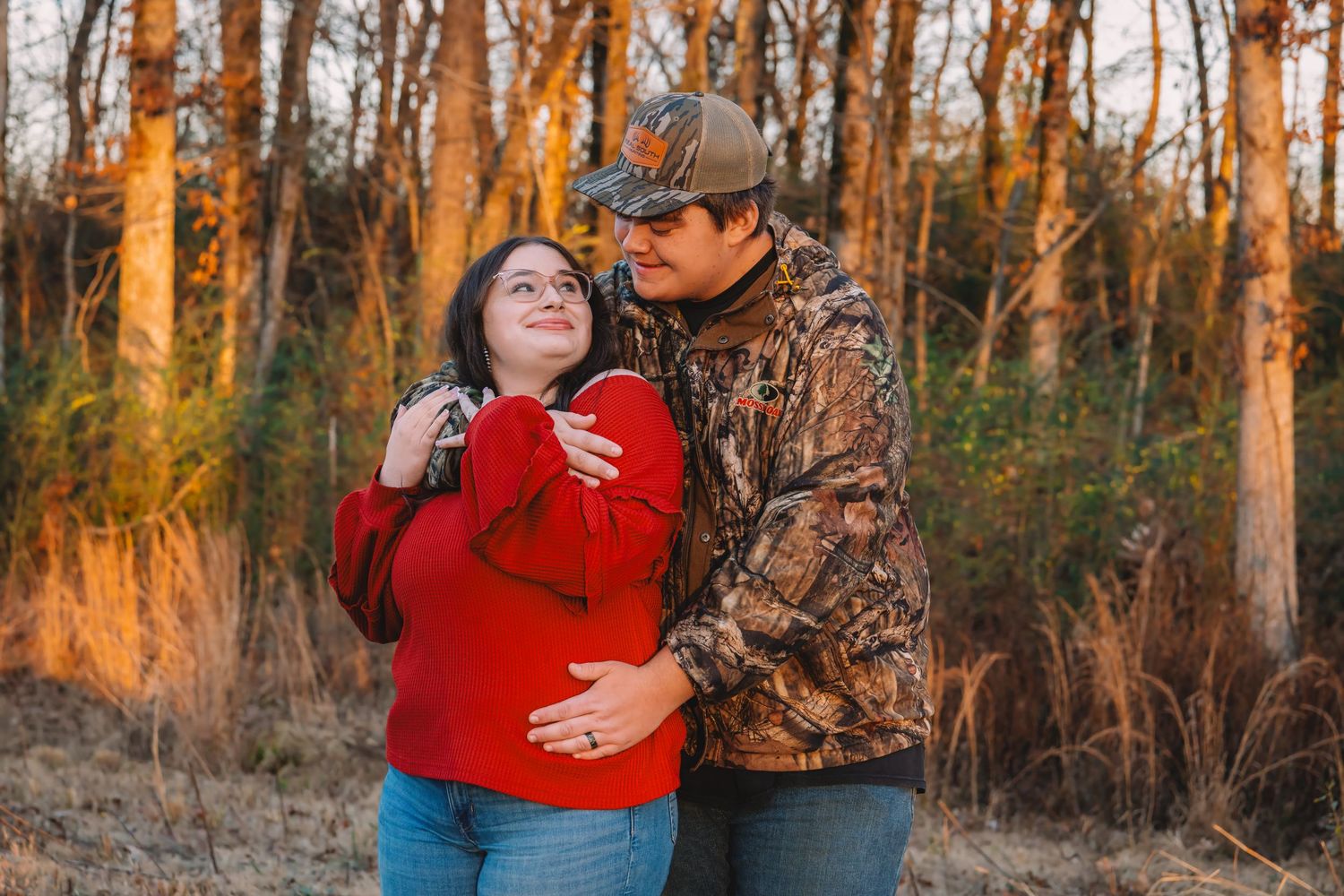 A couple in autumn forest wearing red and camo clothing embraces during golden hour sunset lighting.