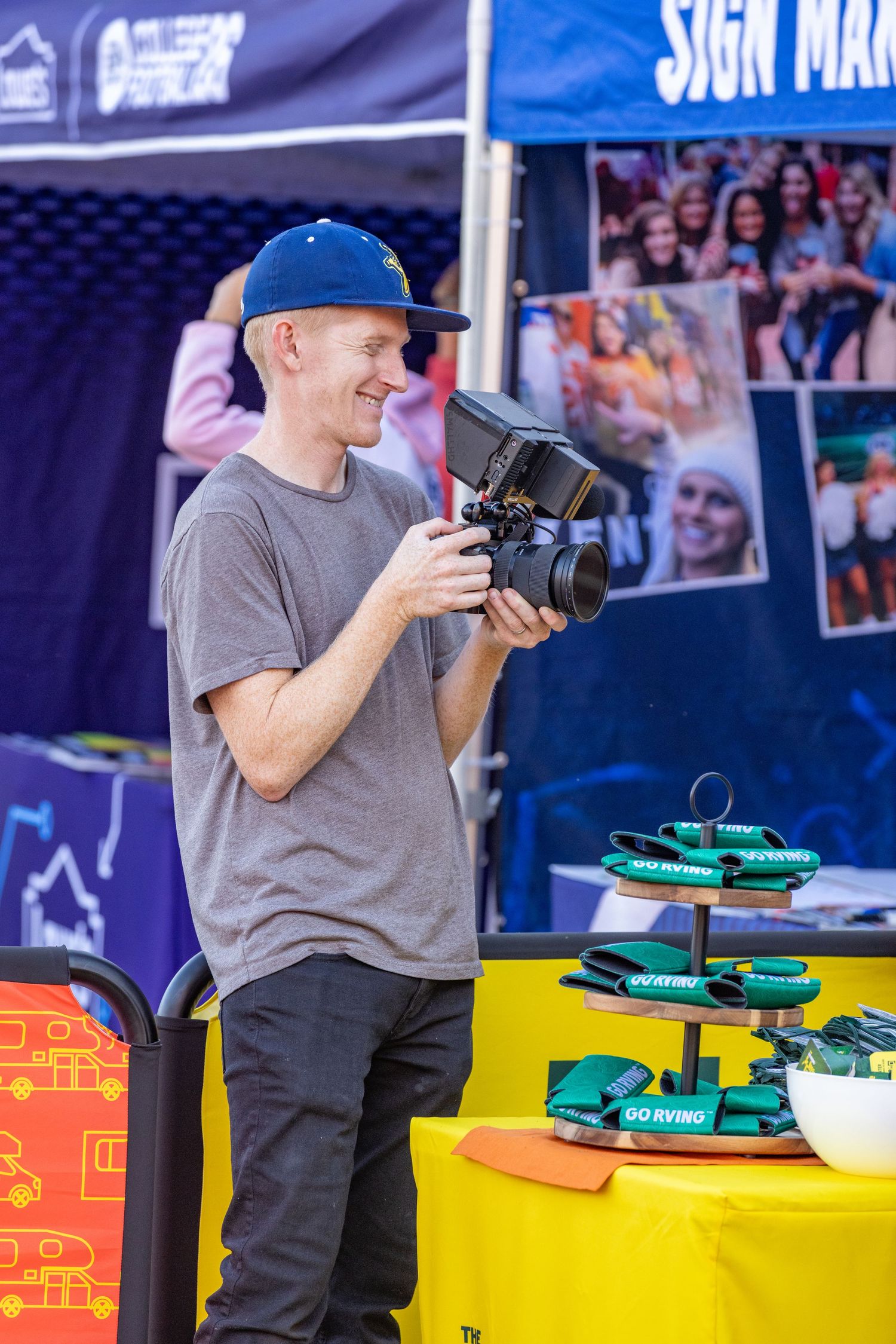 Person in gray shirt and baseball cap interacts with items displayed on yellow table at outdoor event setup.
