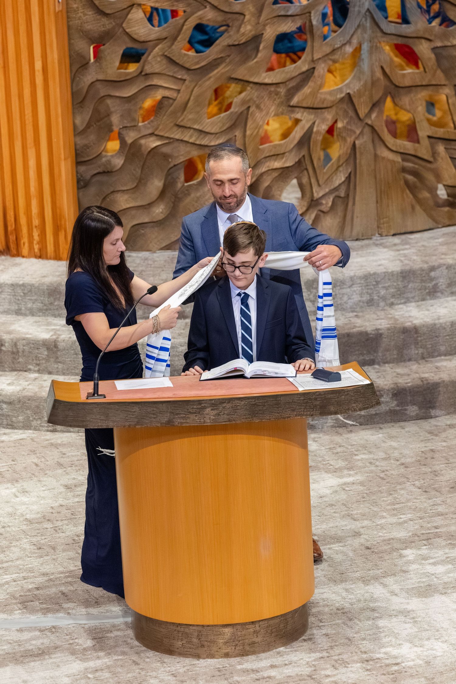 Young boy reads from Torah at bar mitzvah ceremony, flanked by two adults holding tallit at synagogue bimah.