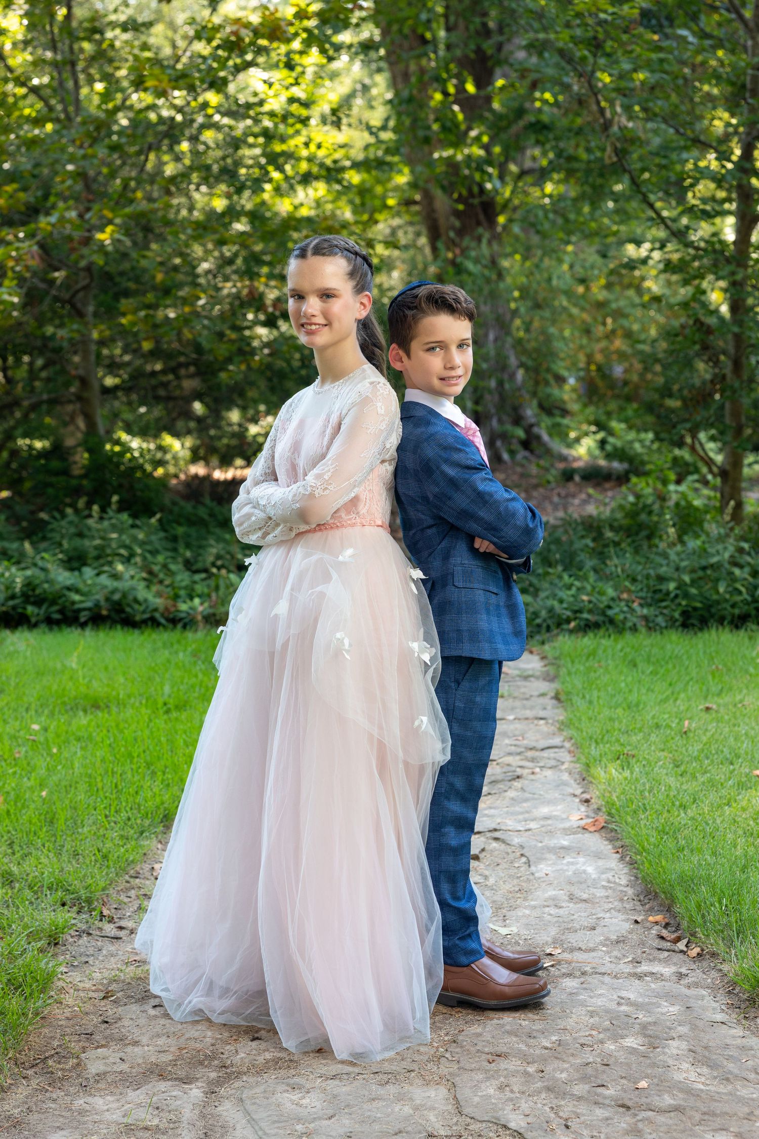 A girl in a pink tulle gown and boy in blue suit stand back-to-back on a stone path in a lush garden.