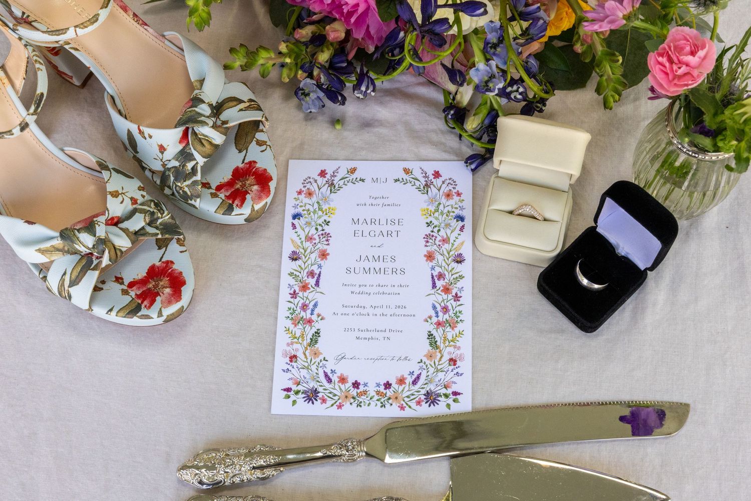 Floral wedding flat lay featuring embroidered heels, wildflower invitation, cake knife, ring boxes, and colorful bouquet.