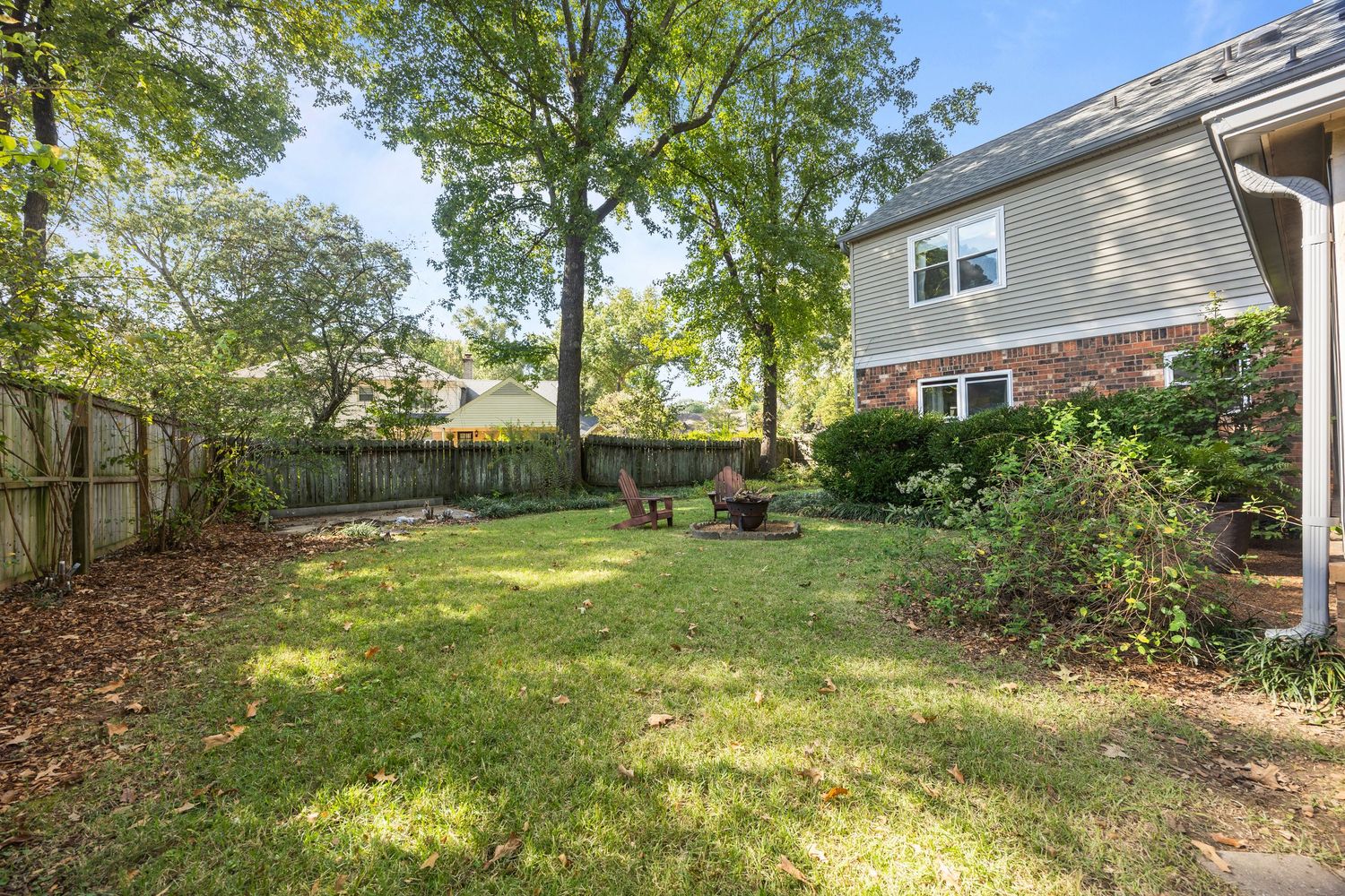 Fenced backyard with green grass, mature trees, and a gray house with brick accents in a residential setting.