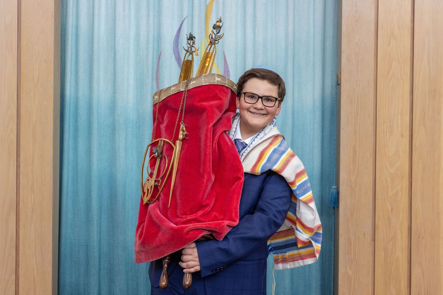 A student hugs a large red cello case while standing against a blue and wooden paneled wall.