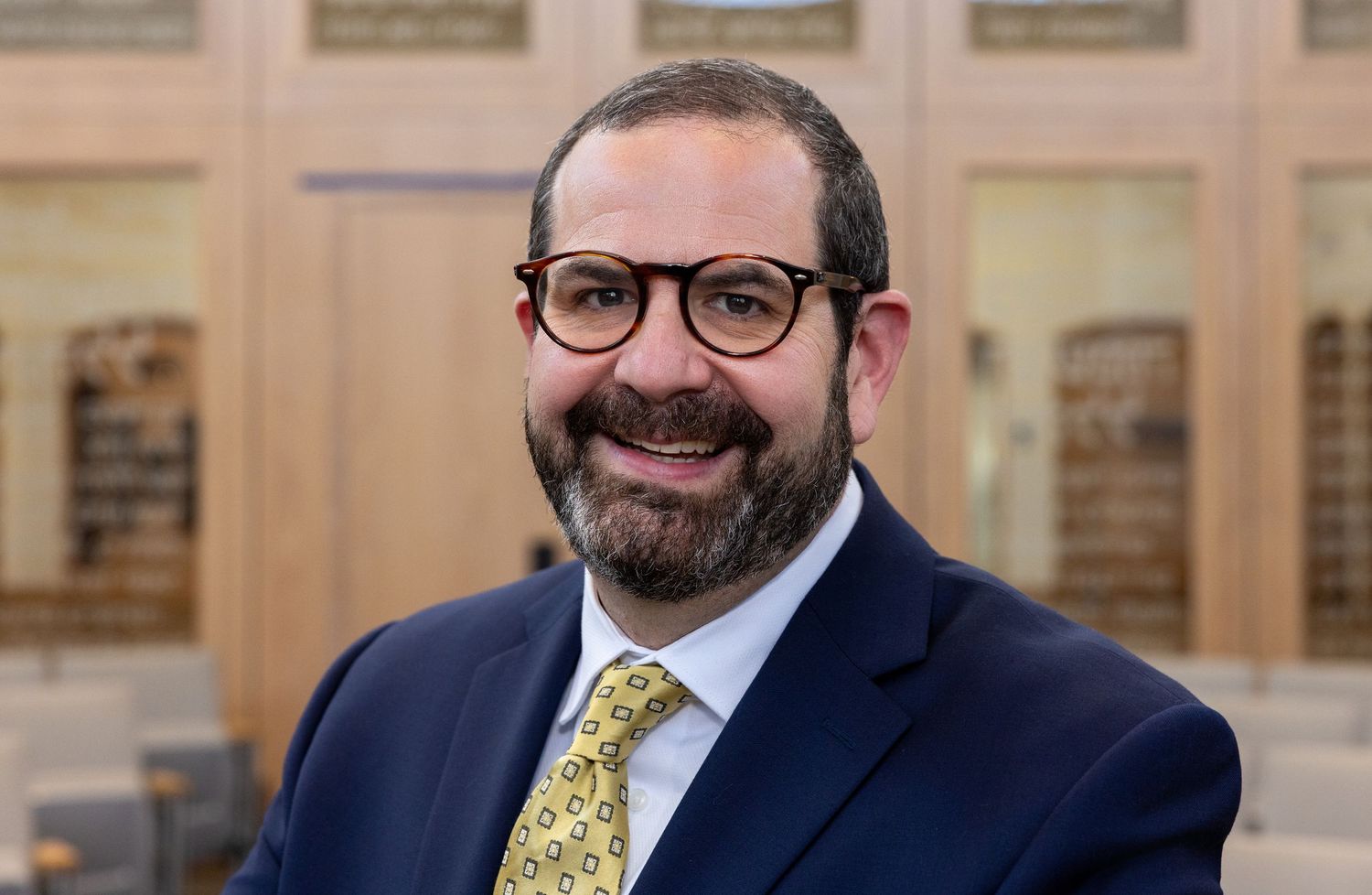 Professional man in navy suit and gold tie with glasses, smiling warmly in an elegant library setting.