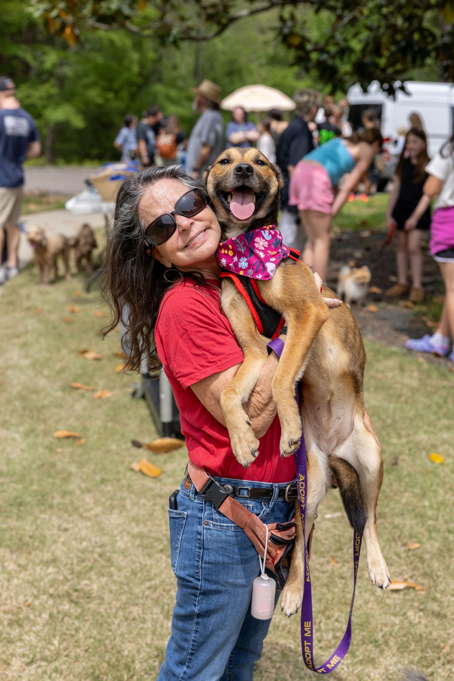Woman in red shirt hugging a happy dog wearing a pink bandana at an outdoor pet adoption event.