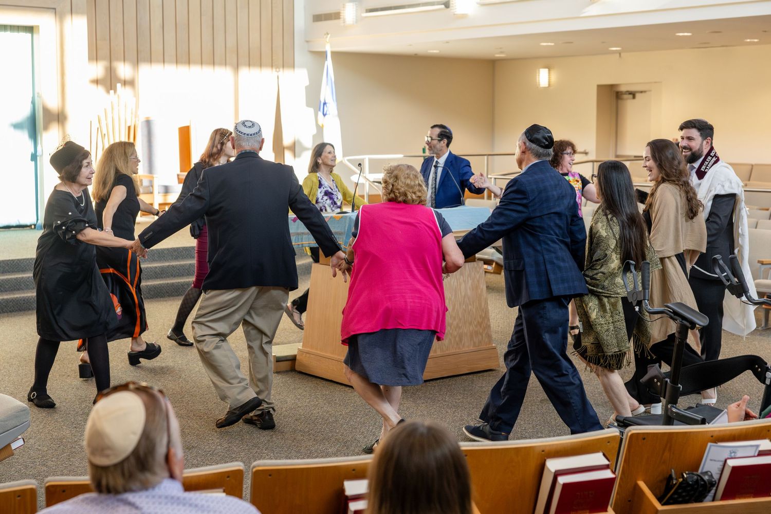 Guests dance joyfully around a wooden box during a Jewish celebration inside a synagogue.