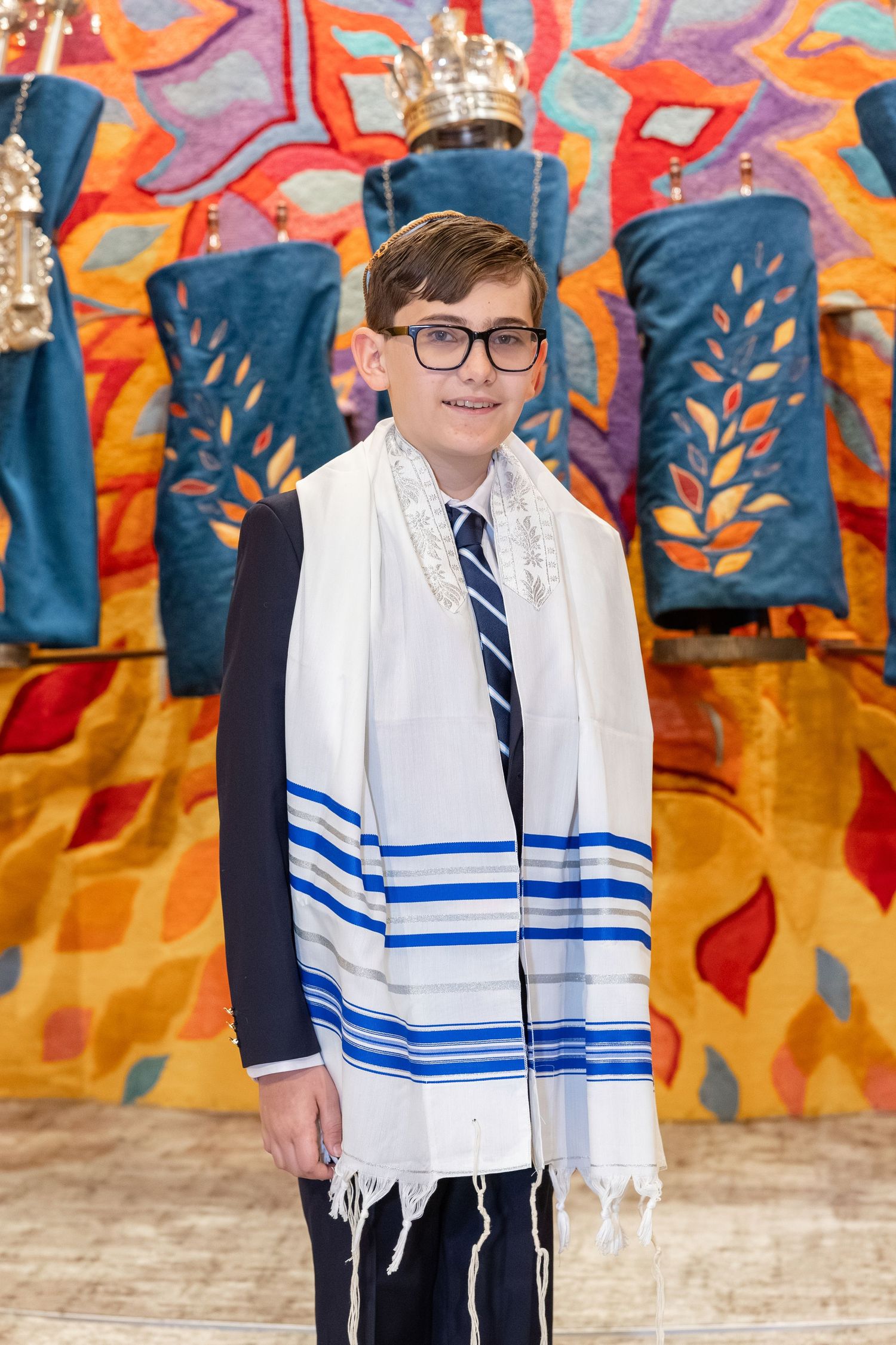 Young boy in tallit and glasses smiling at his Bar Mitzvah ceremony with Torah scrolls in background.