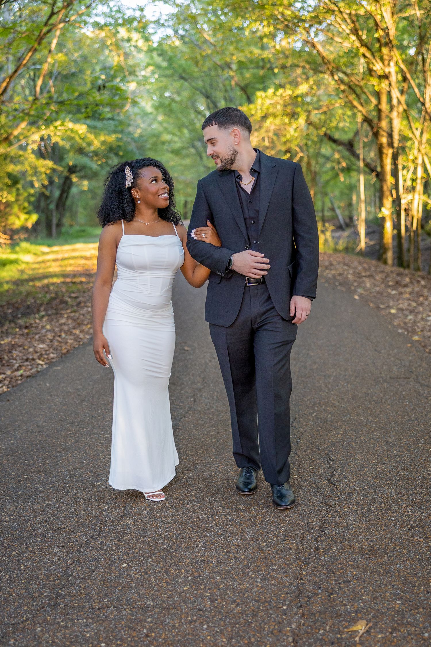 Couple in formal attire strolling along a scenic wooded path during sunset.