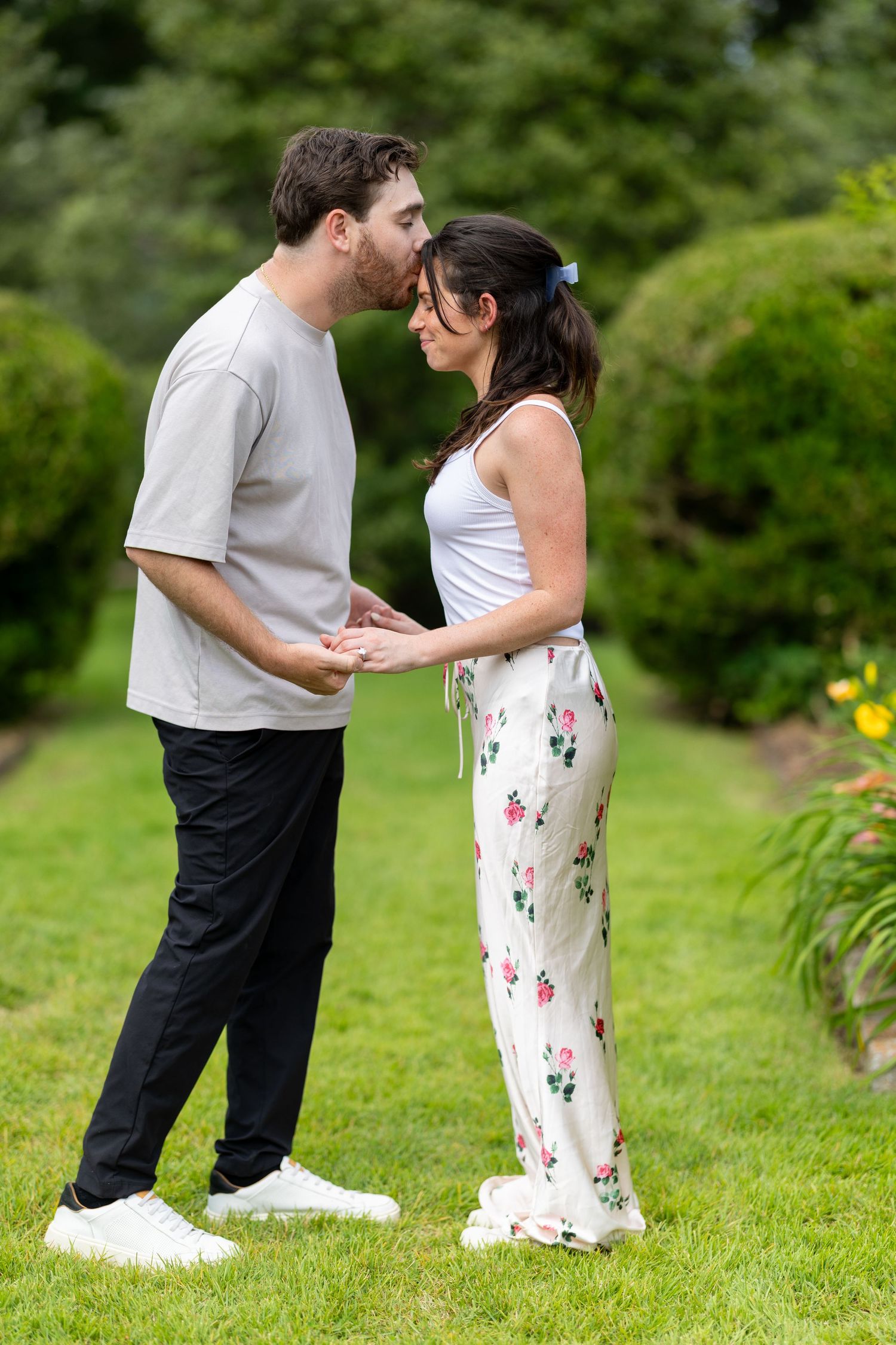 Couple holding hands and sharing a tender forehead kiss in a lush green garden park.