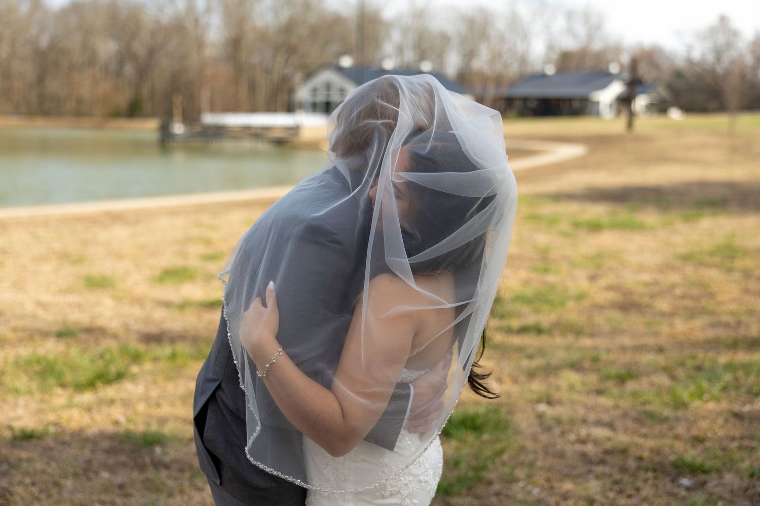 A couple shares a tender moment outdoors with the bride's veil flowing in the breeze against a pastoral background.