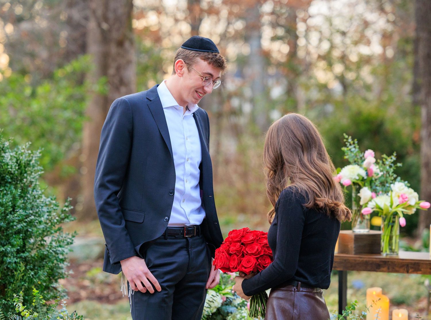 A couple shares a romantic moment outdoors with red roses and floral arrangements in a wooded setting at sunset.