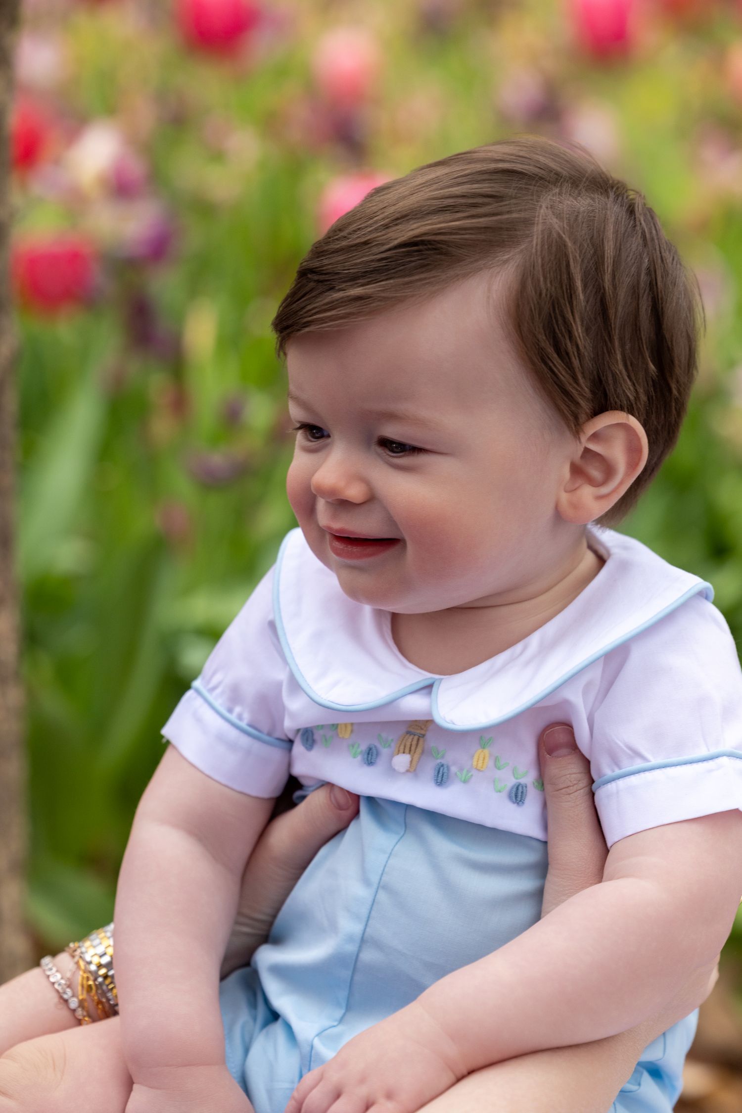 A baby in a blue and white outfit smiles while sitting in a garden with pink flowers visible behind.