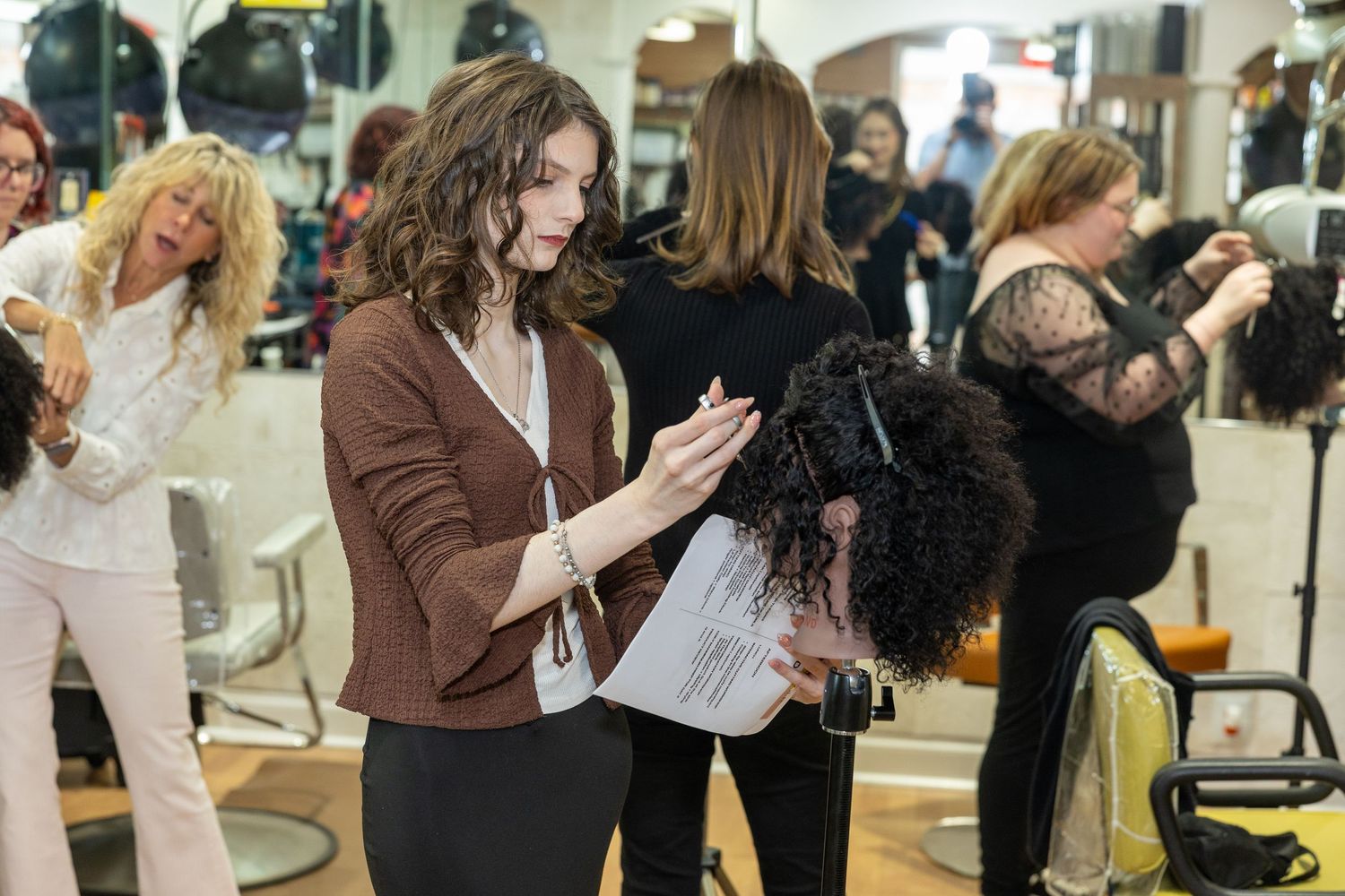 Hair stylist reviewing notes with client in a busy professional salon with stylists working in background.