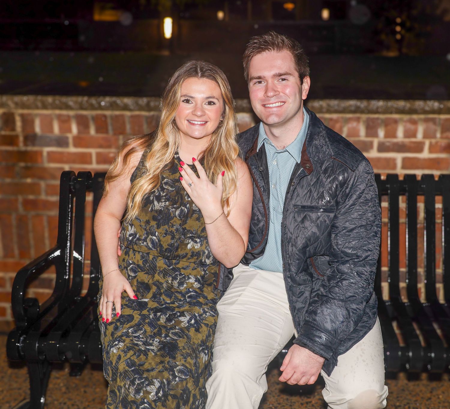 Happy newly engaged couple sitting on bench showing off ring at night.