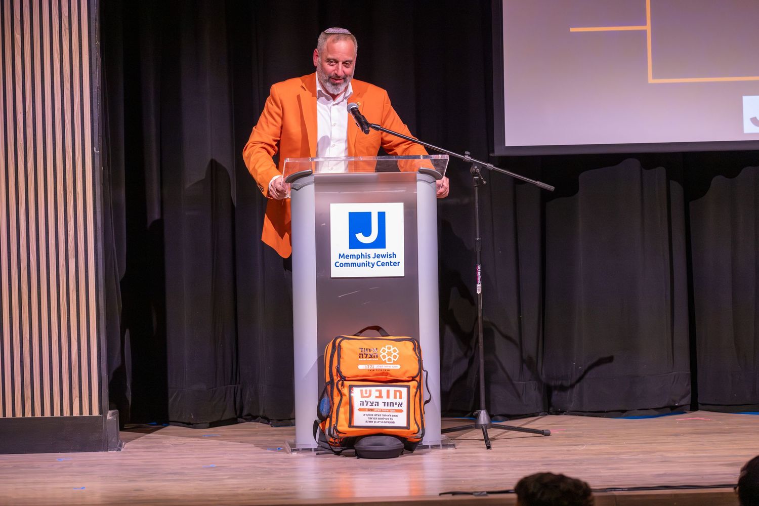 Speaker in orange jacket stands at podium during presentation.