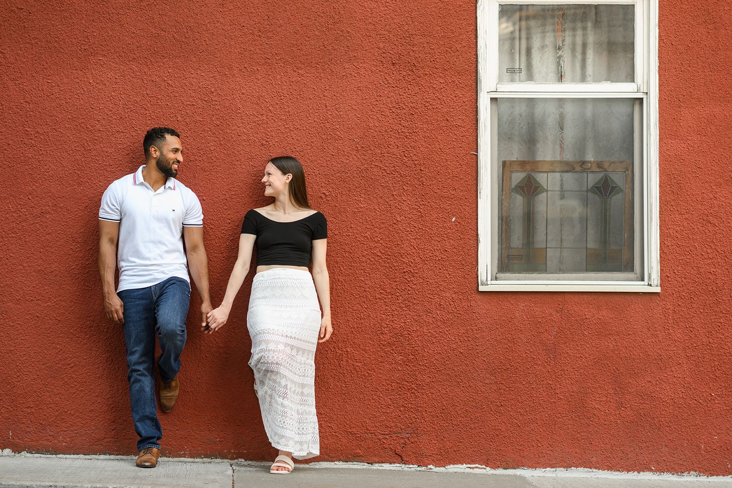 Couple holding hands and smiling at each other against a red wall outdoors.