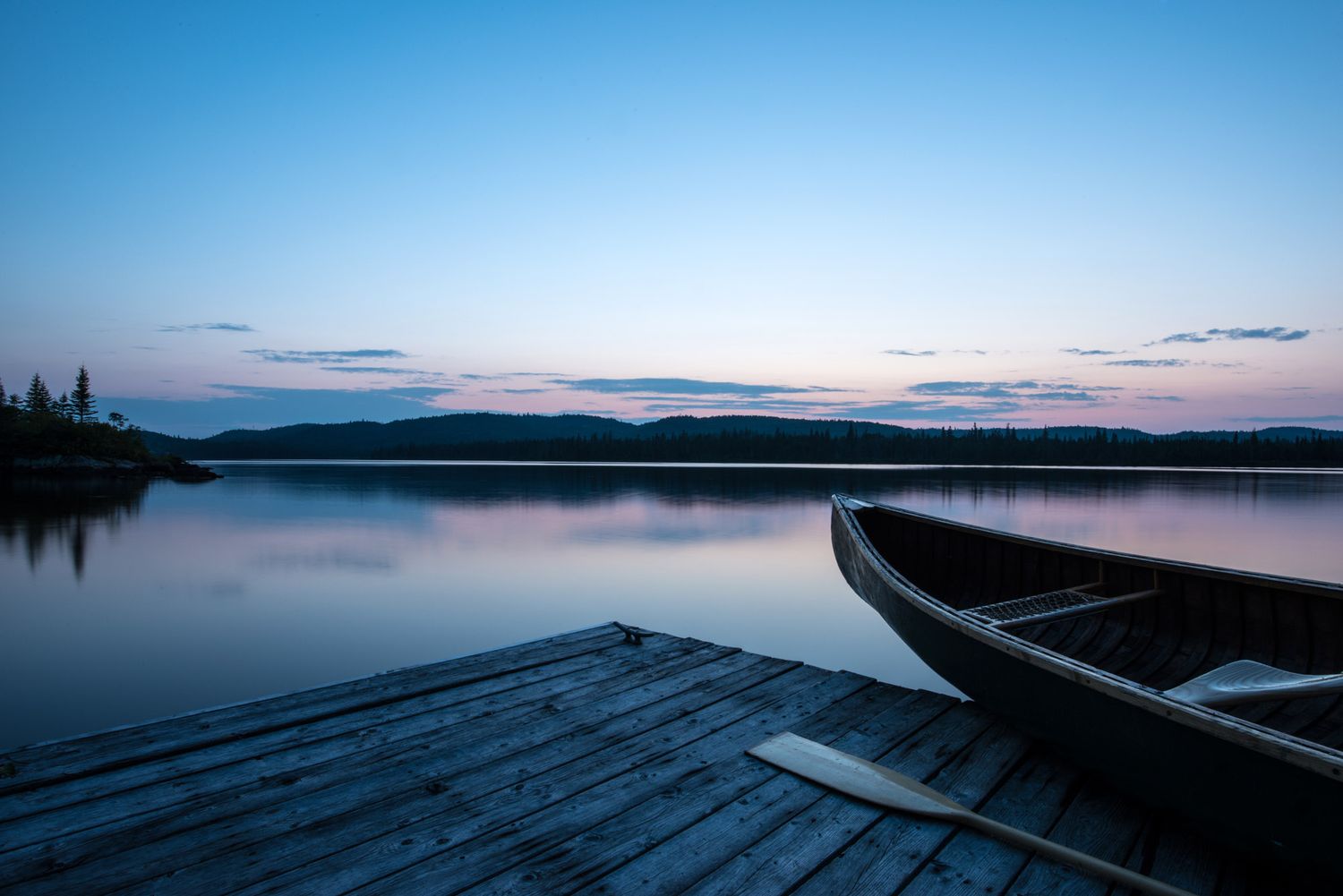 A wooden dock and canoe rest beside a calm lake at dusk, with mountains reflecting in the still water.