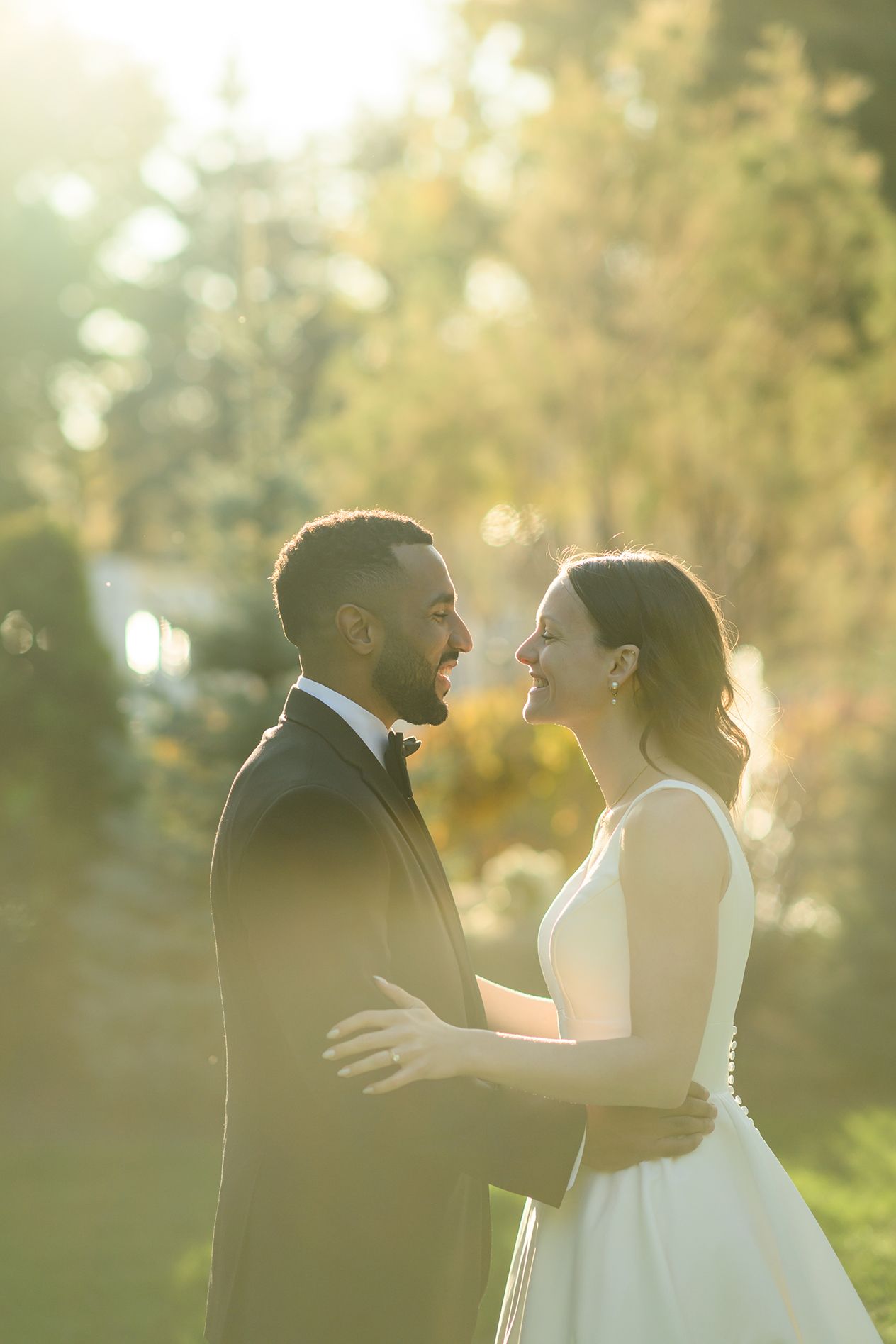 A bride and groom share a joyful moment outdoors, embracing in golden sunlight with lush green trees in the background.
