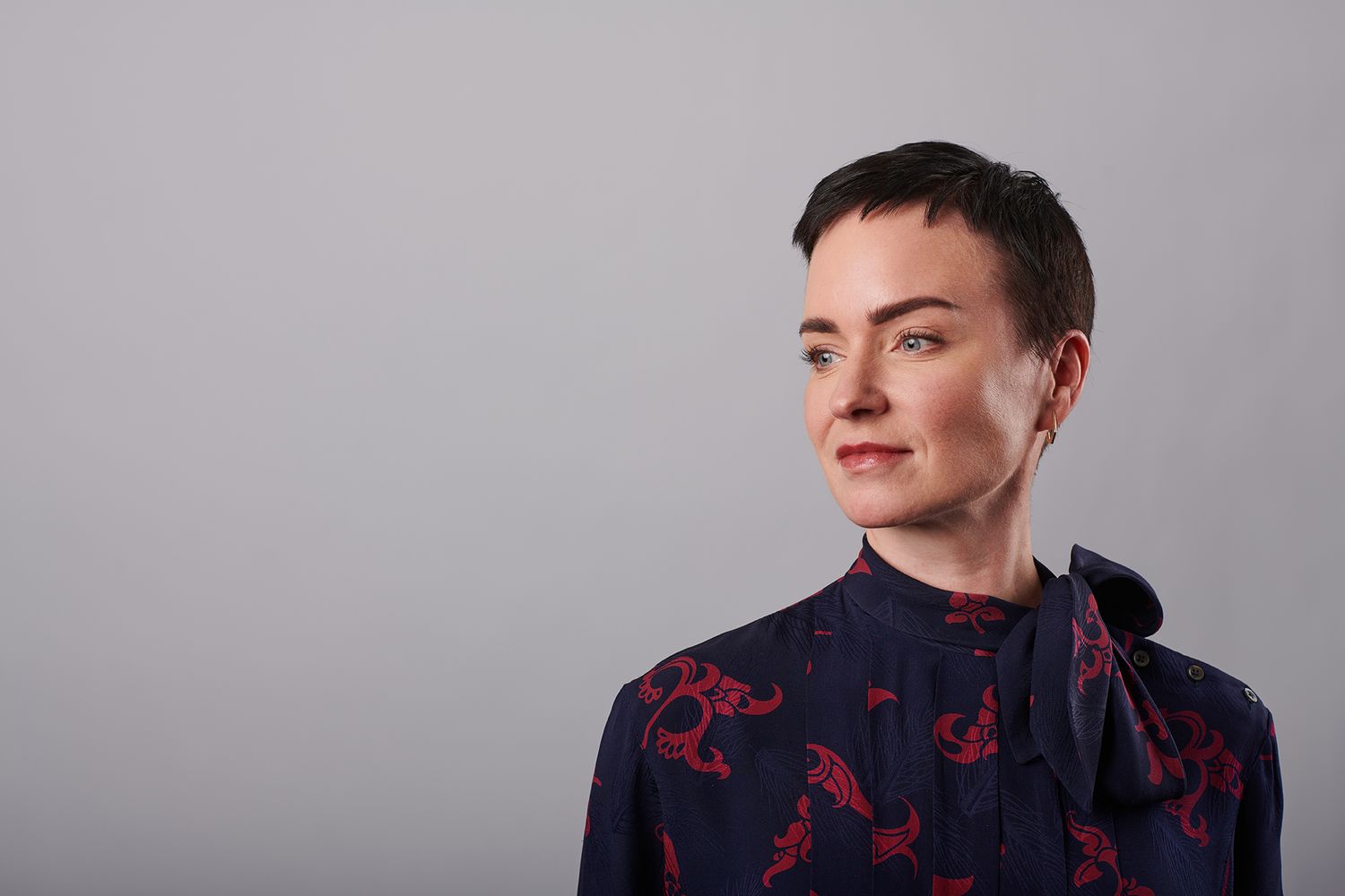 Woman with short dark hair wearing a navy floral blouse with bow tie, posing against a gray background.