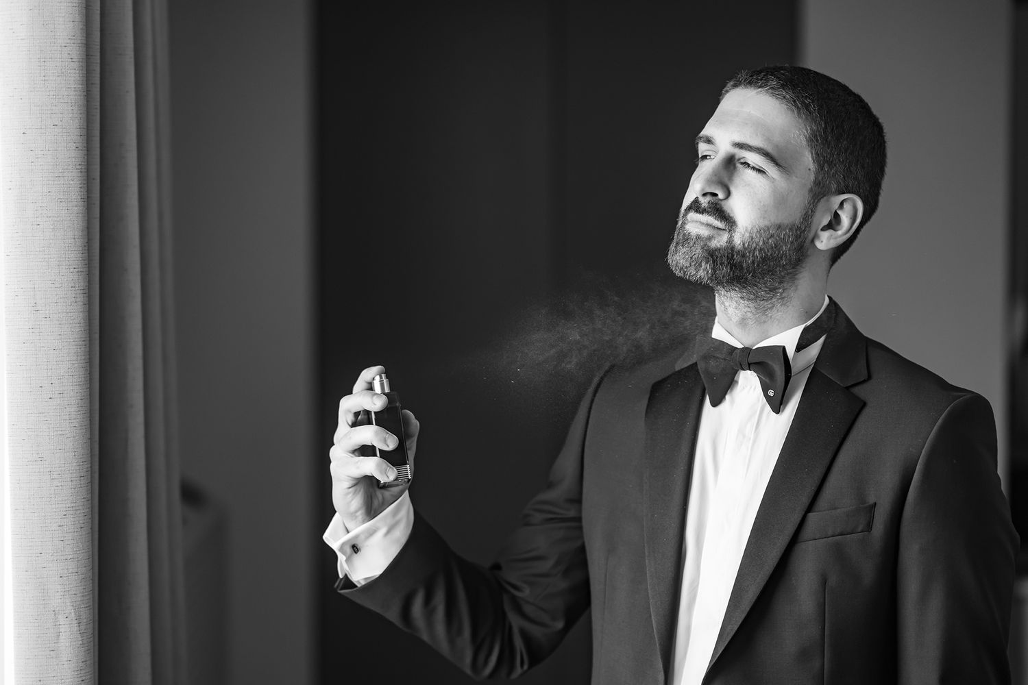 Man in tuxedo and bow tie spraying cologne in black and white photo.