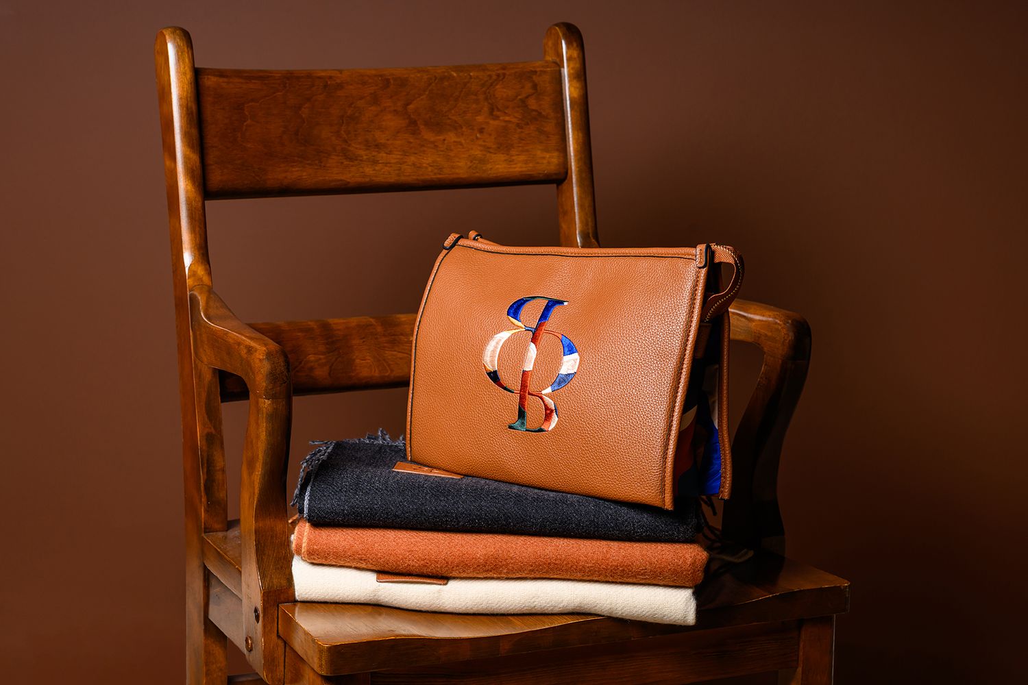 Brown leather crossbody bag with monogram on folded denim and fabric on a wooden chair.