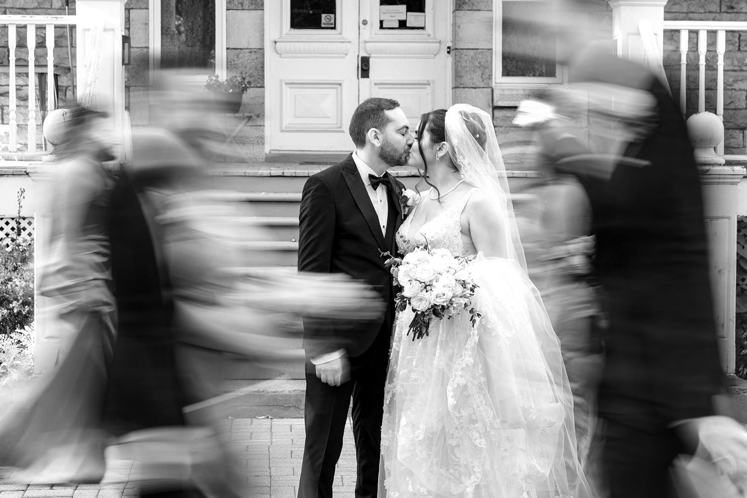 Black and white wedding photo of bride and groom kissing while wedding party moves around them in blur.