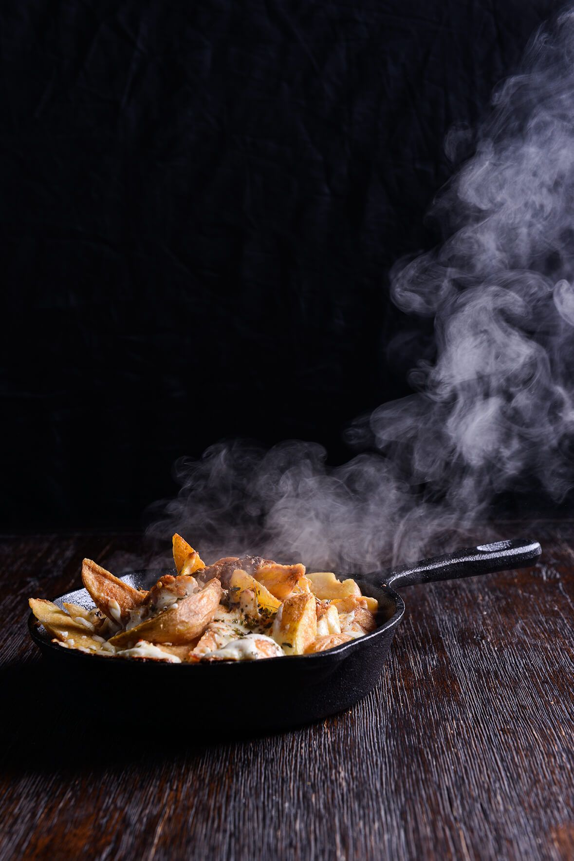 Steaming cast iron skillet filled with roasted potato wedges and melted cheese on a rustic wooden table against dark background.