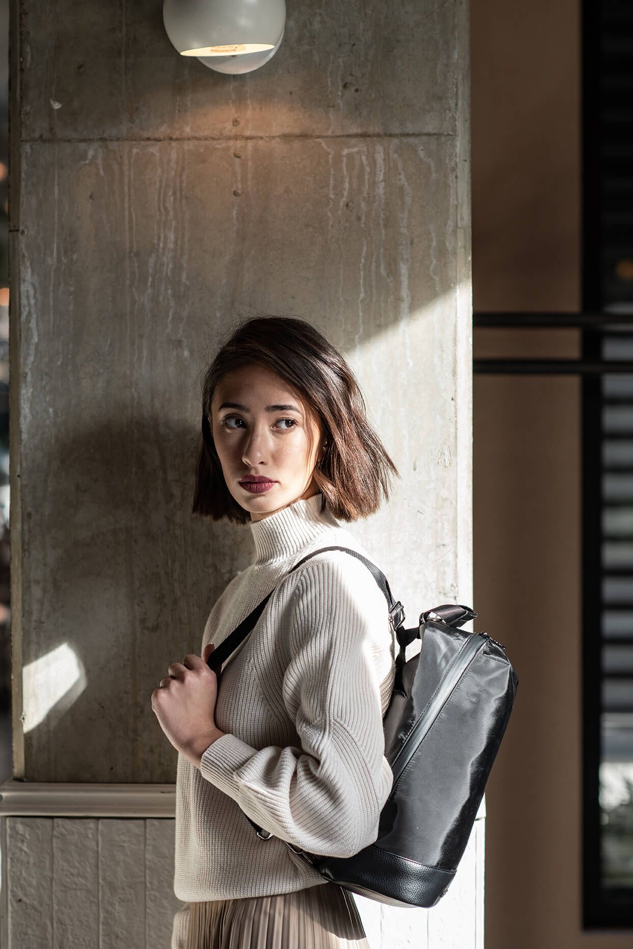 Young woman with short hair wearing a cream ribbed turtleneck sweater, carrying a black leather backpack indoors.
