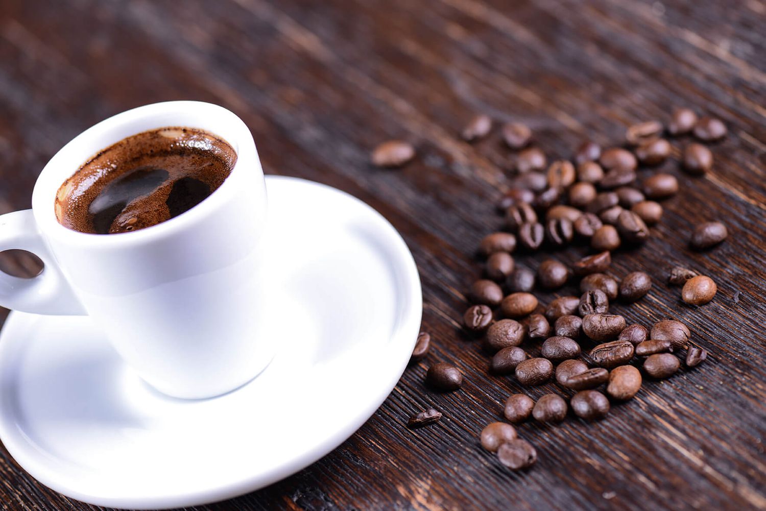 White espresso cup on saucer with dark coffee beside scattered roasted coffee beans on rustic wooden table.