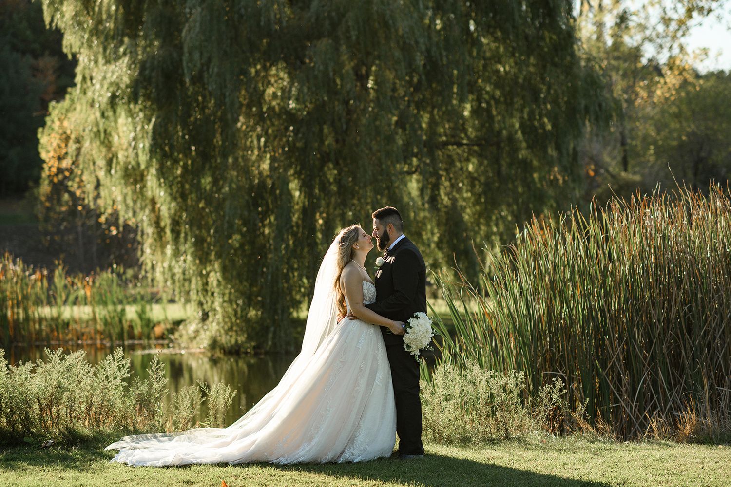 Bride and groom share a romantic moment by a pond under a weeping willow tree in a lush garden setting.