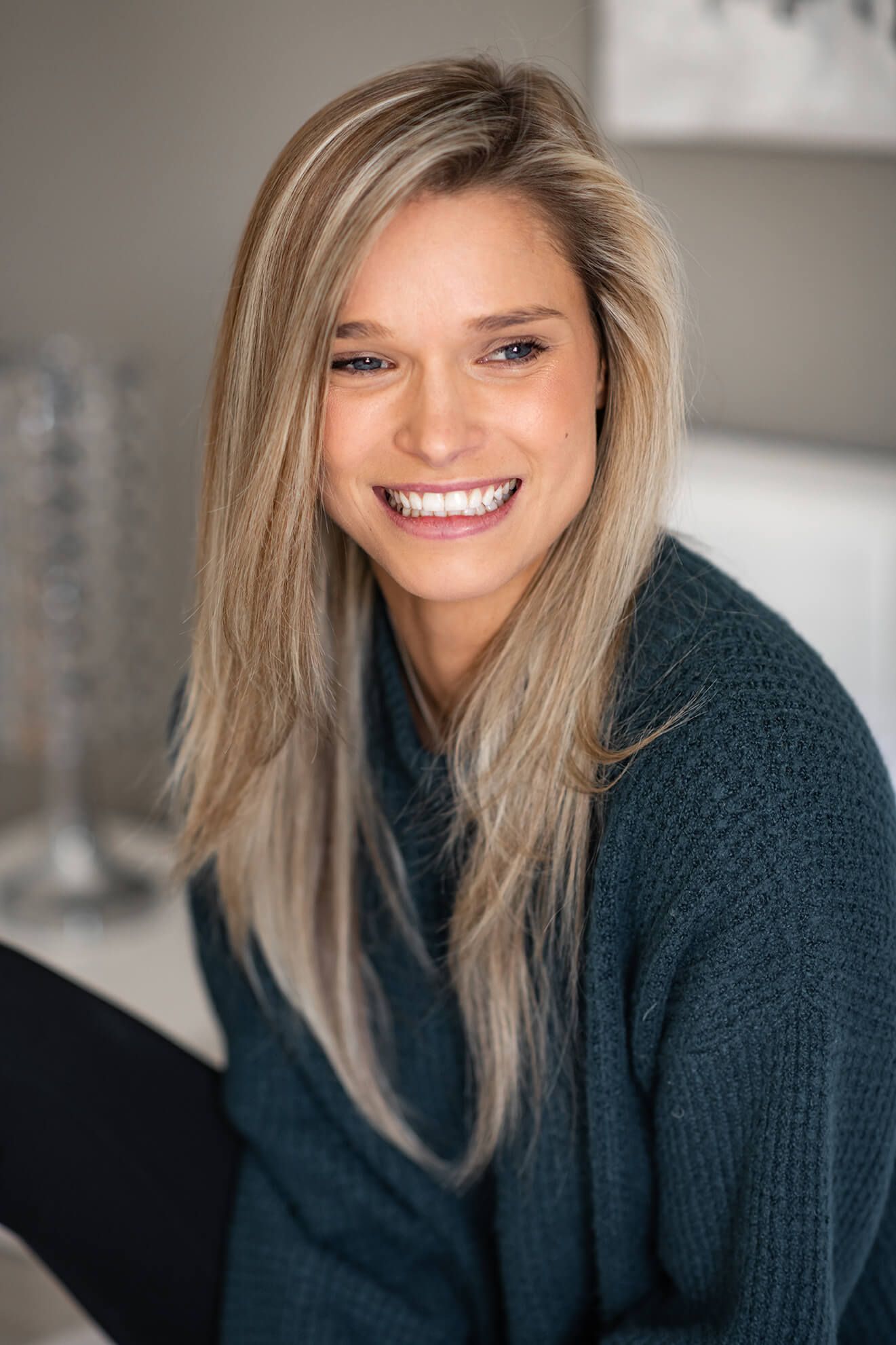 Smiling blonde woman in a teal knit sweater with long straight hair poses indoors in a bright modern setting.