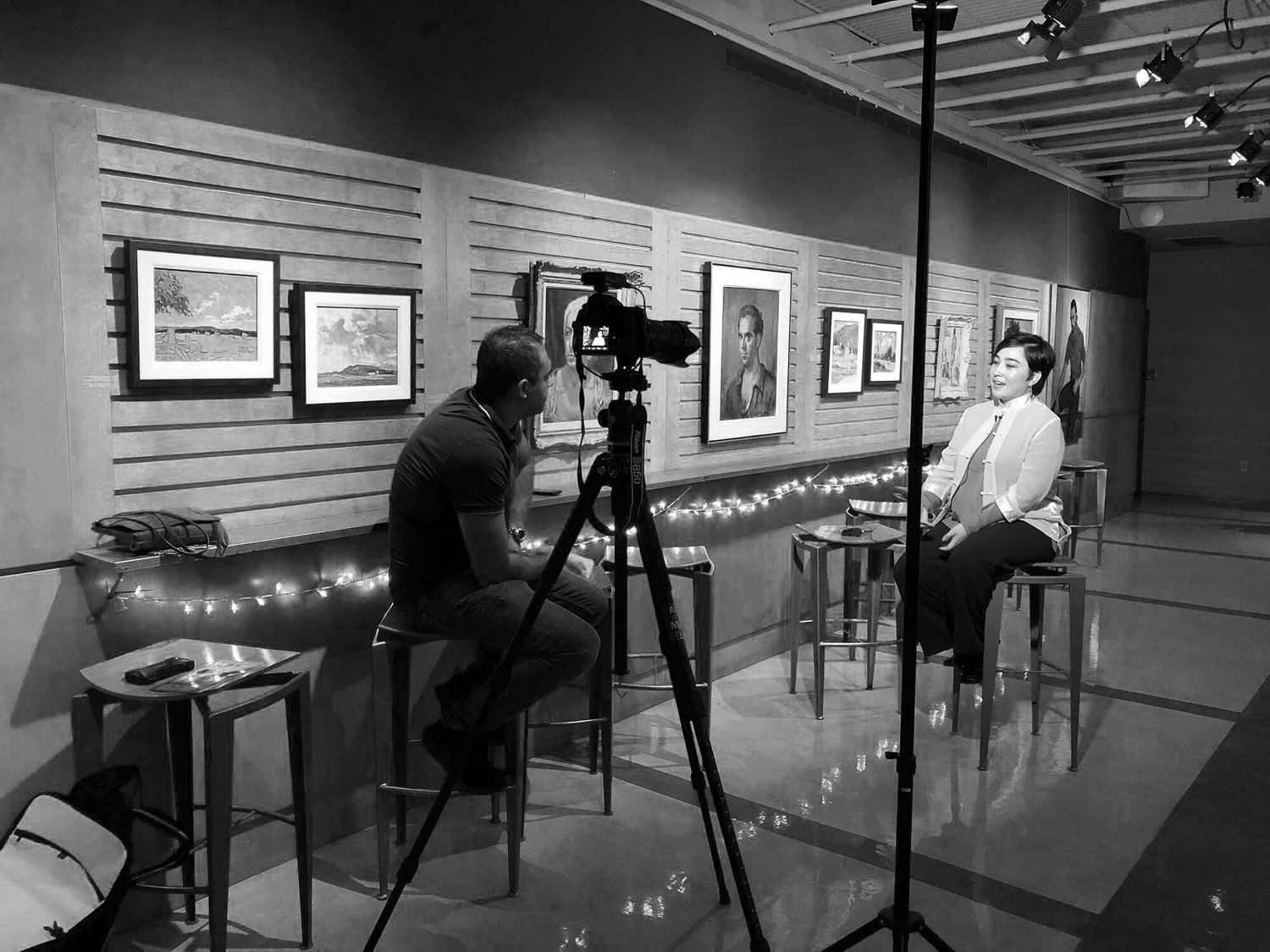 Black and white photo of a videographer filming an interview in an art gallery with framed artwork on walls.