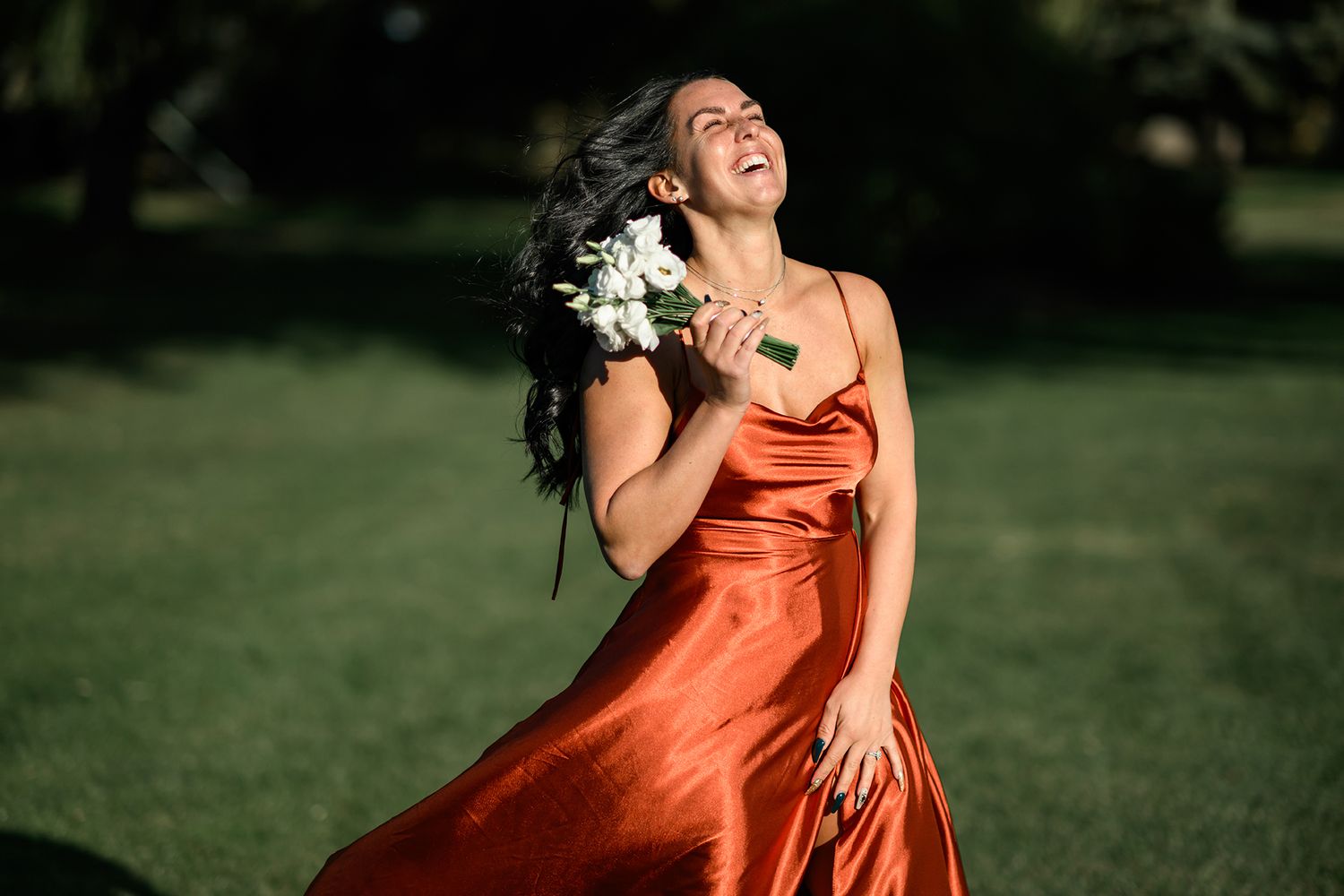 Woman in rust satin slip dress laughing joyfully outdoors, holding white bouquet, hair flowing in breeze.