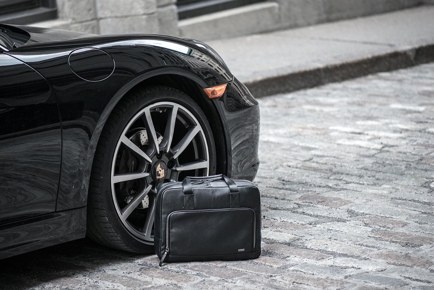 Black Porsche sports car with a sleek leather travel bag placed beside the rear wheel on cobblestone pavement.